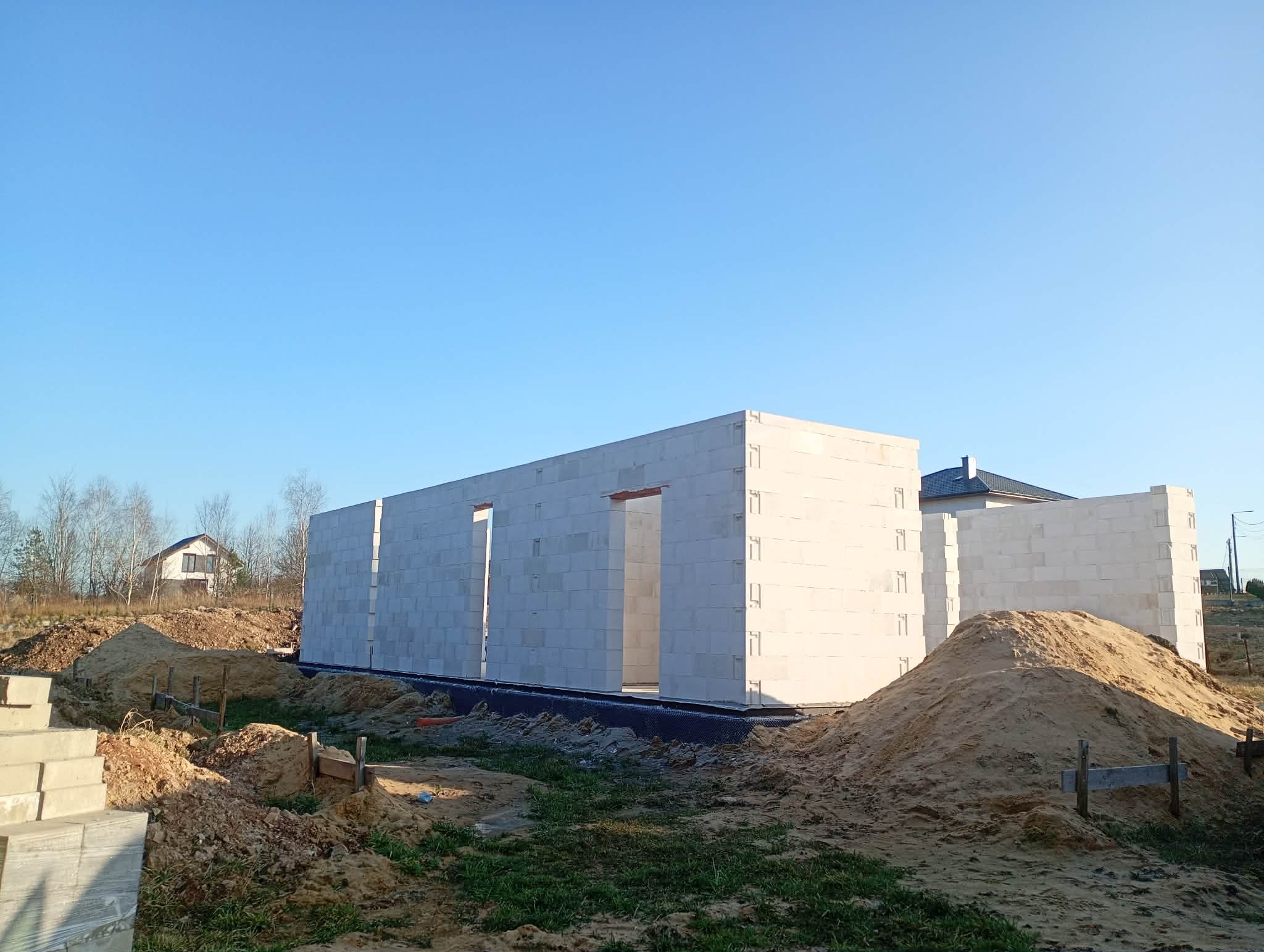Wznoszone ściany domu z betonu komórkowego na fundamencie, widoczne otwory okienne i drzwiowe, hałdy piasku, w tle dom jednorodzinny i błękitne niebo.