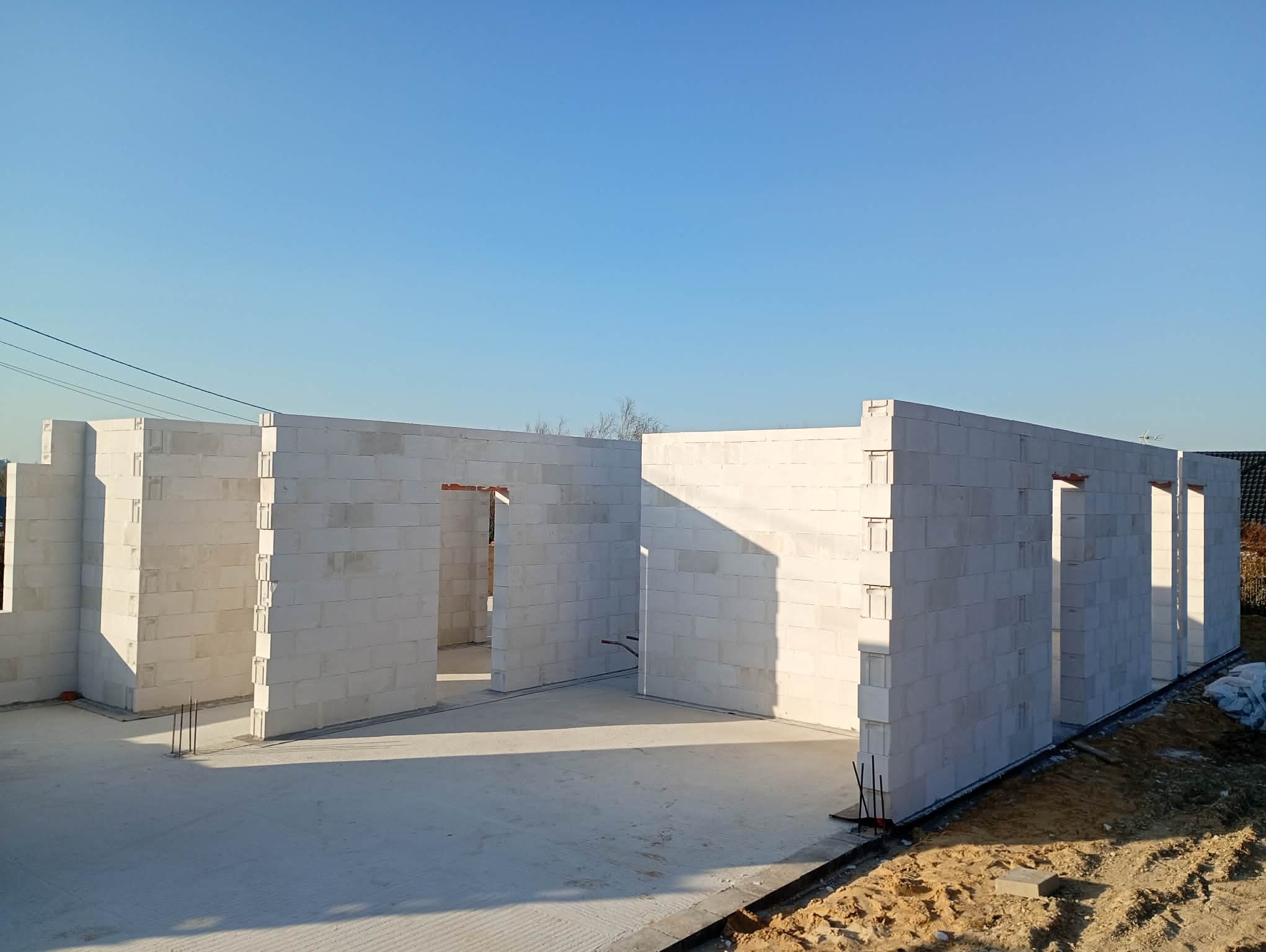 Wnętrze domu w trakcie budowy, ściany z białych bloczków, widoczne otwory okienne i drzwiowe, betonowa podłoga, błękitne niebo w tle. Surowy stan budynku.