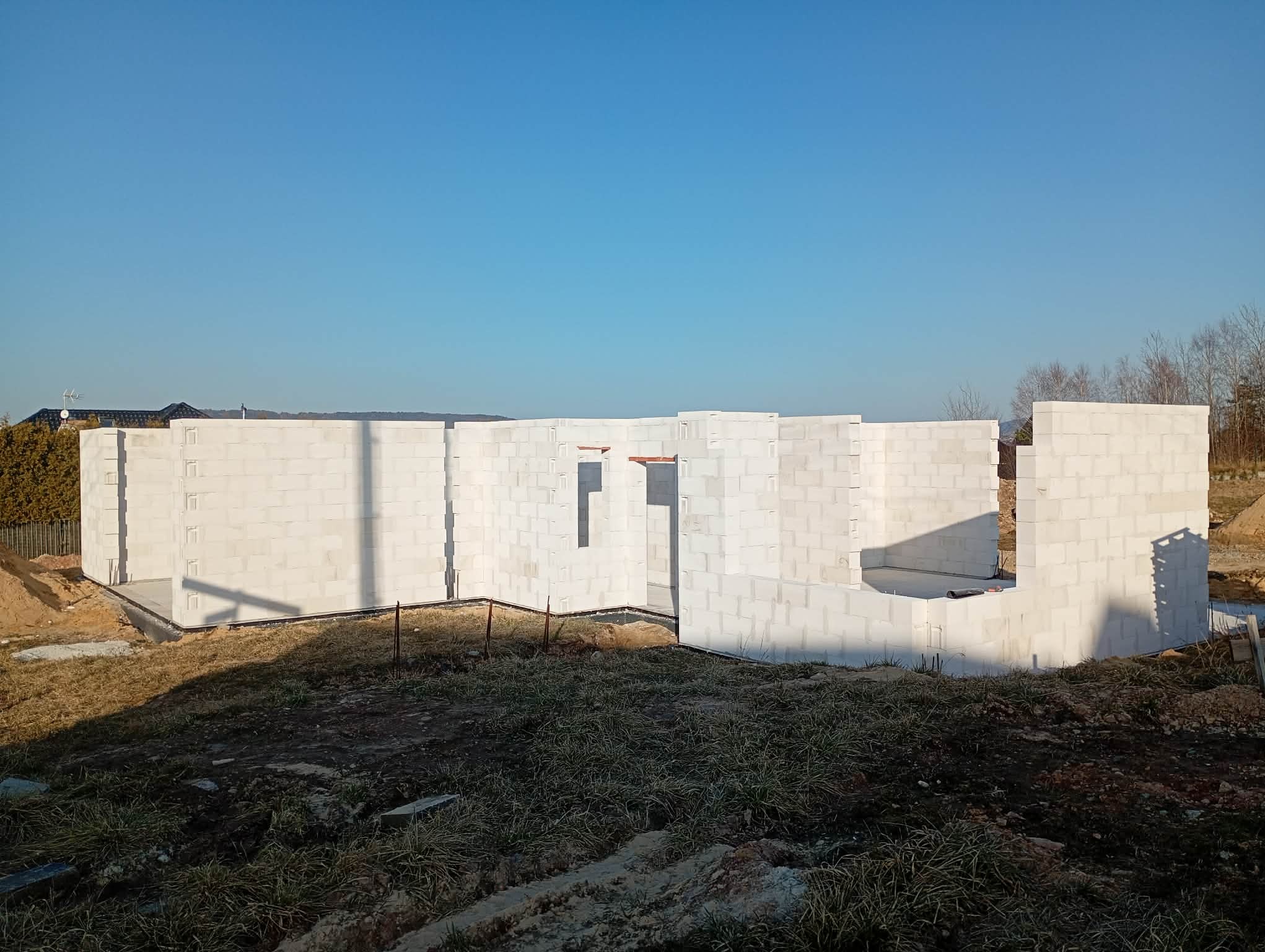 Wznoszenie ścian domu z białych bloczków betonu komórkowego. Widoczne otwory okienne i drzwiowe. Fundamenty i początek ścian na tle błękitnego nieba.