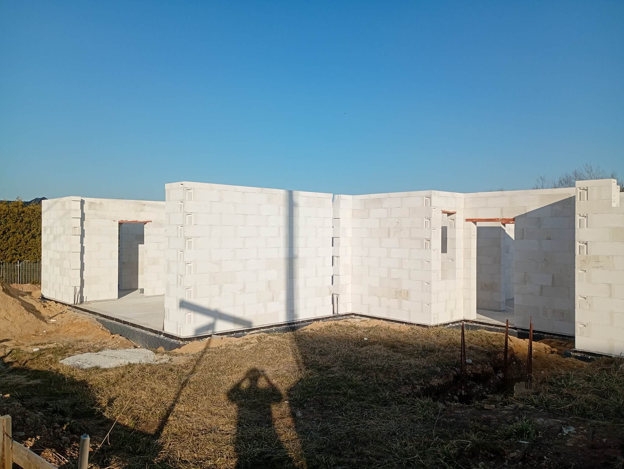 Wznoszenie ścian parteru z białych bloczków betonu komórkowego na fundamencie. Widoczny cień osoby fotografującej. Błękitne niebo w tle.