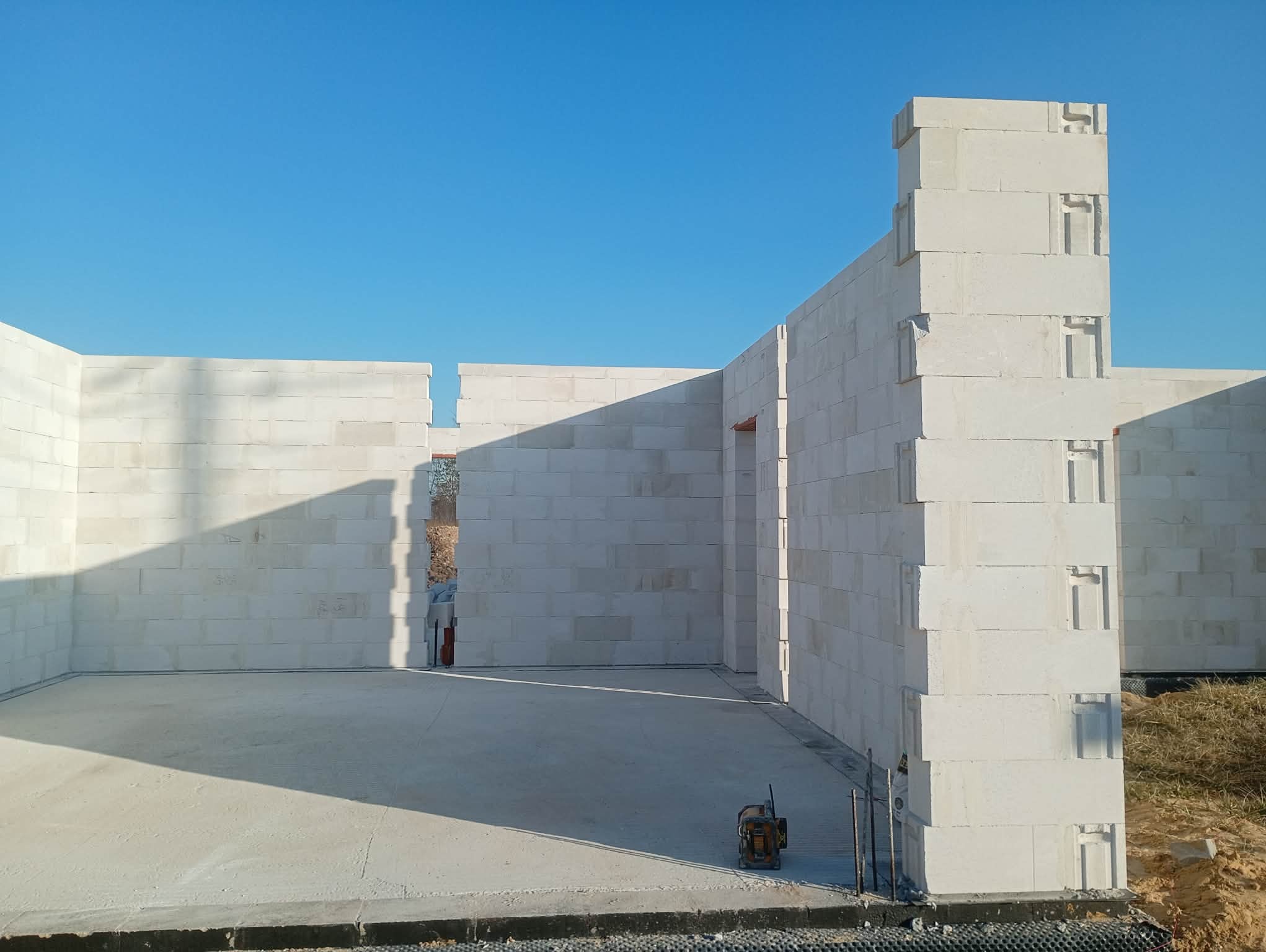 Wnętrze domu w trakcie budowy z białych bloczków, widoczne ściany i wylewka, narzędzie budowlane w rogu. Słoneczny dzień, błękitne niebo.