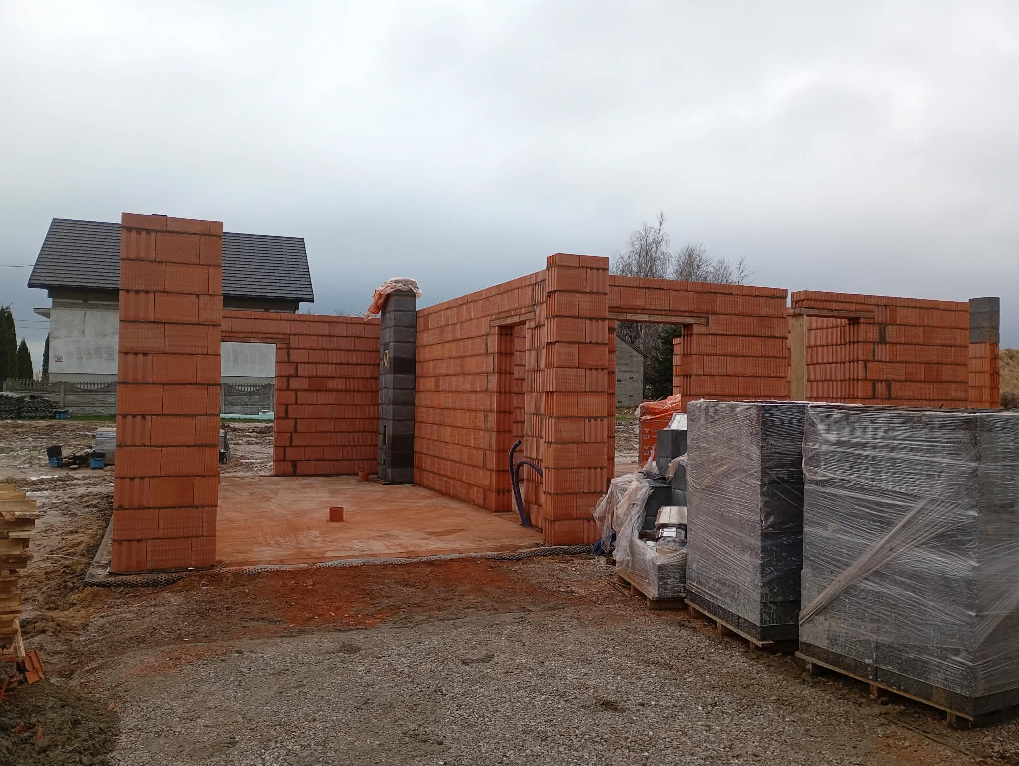 Wznoszenie ścian parteru z czerwonej cegły ceramicznej. Widoczne otwory okienne i drzwiowe. Na paletach zapakowane bloczki. W tle dom z ciemną dachówką.