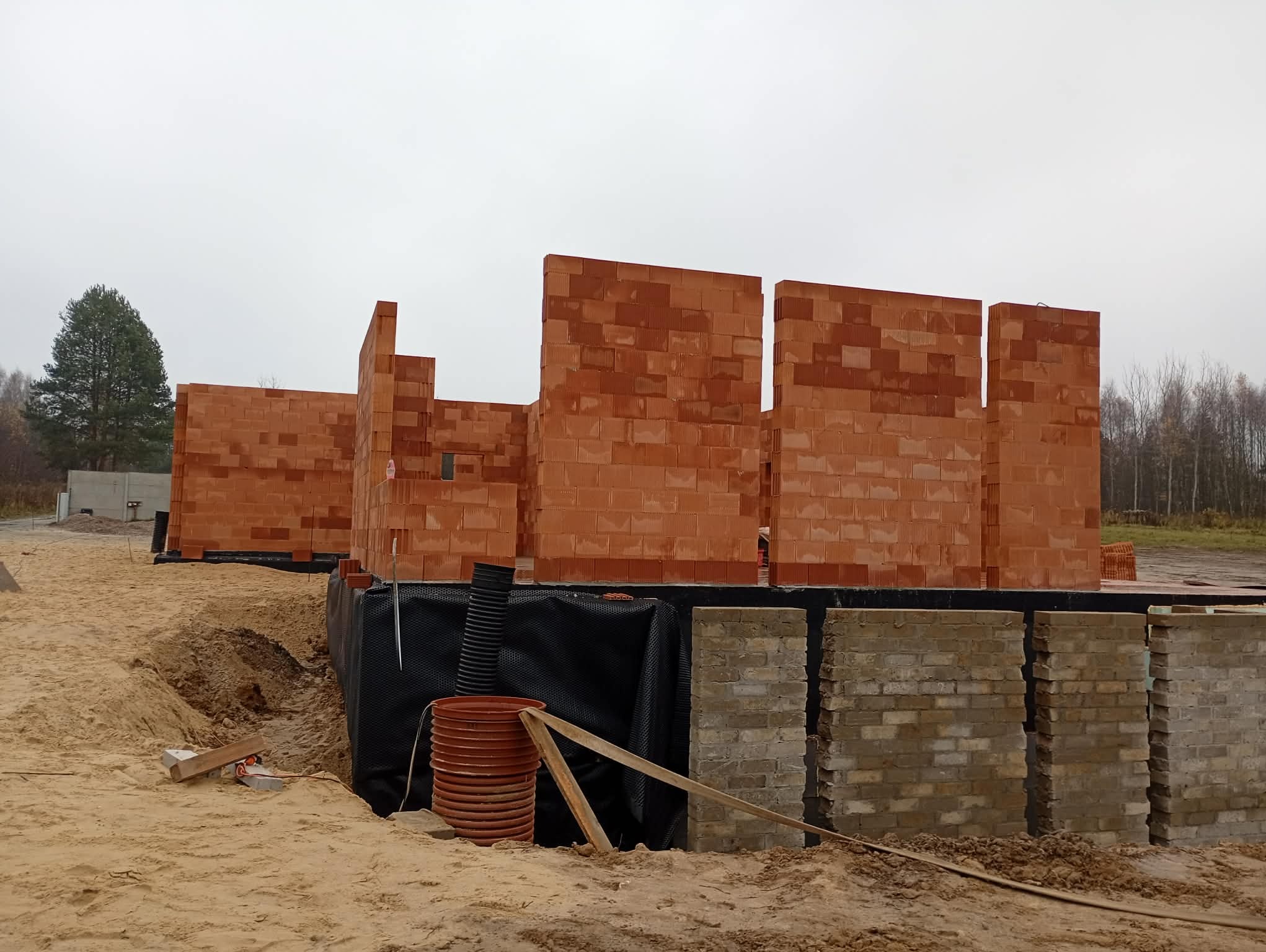 Wznoszone ściany domu z czerwonej cegły na fundamencie z bloczków. Widoczne zabezpieczenia przeciwwilgociowe i elementy szalunku. Prace murarskie w toku, teren budowy.