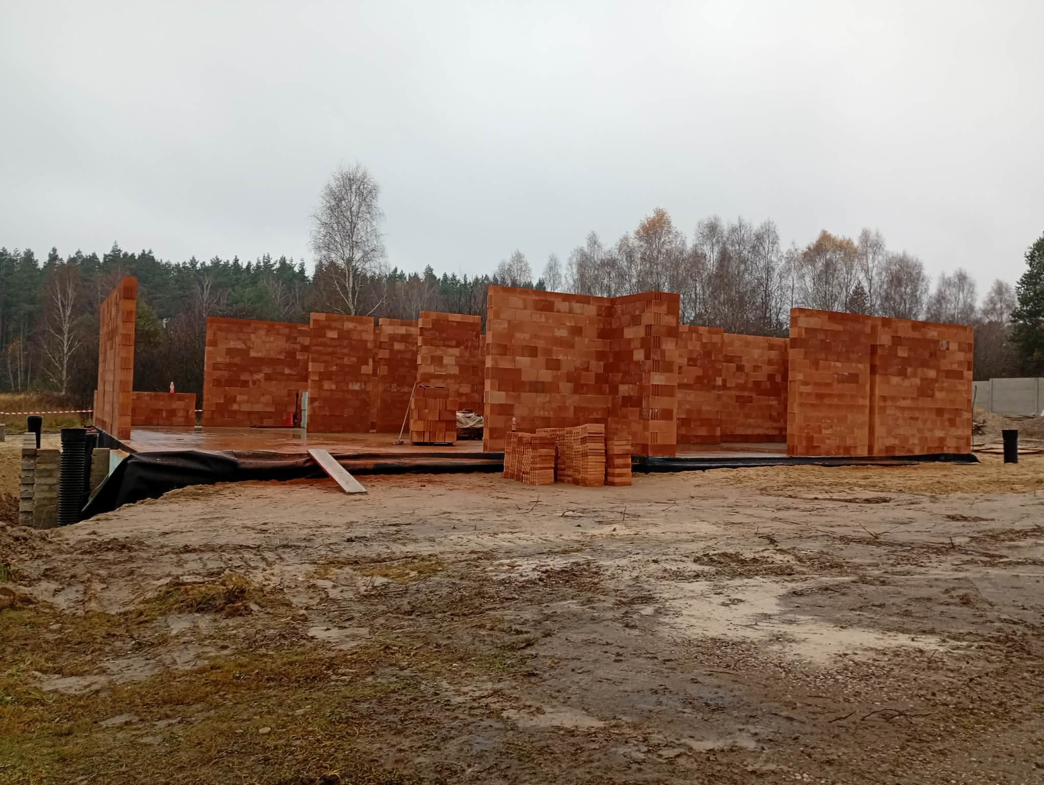 Wznoszone ściany z czerwonej cegły na fundamencie, widoczne z zewnątrz w pochmurny dzień. Częściowo ułożone cegły czekają na użycie. W tle las.