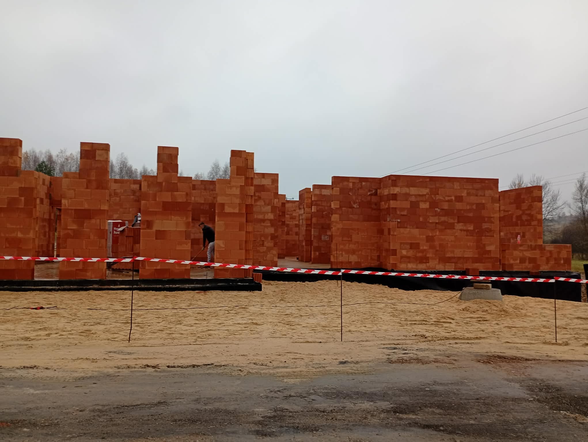Wznoszenie ścian z czerwonej cegły na budowie domu. Widoczny pracownik wyrównujący piasek wewnątrz fundamentów, otoczonych taśmą ostrzegawczą.