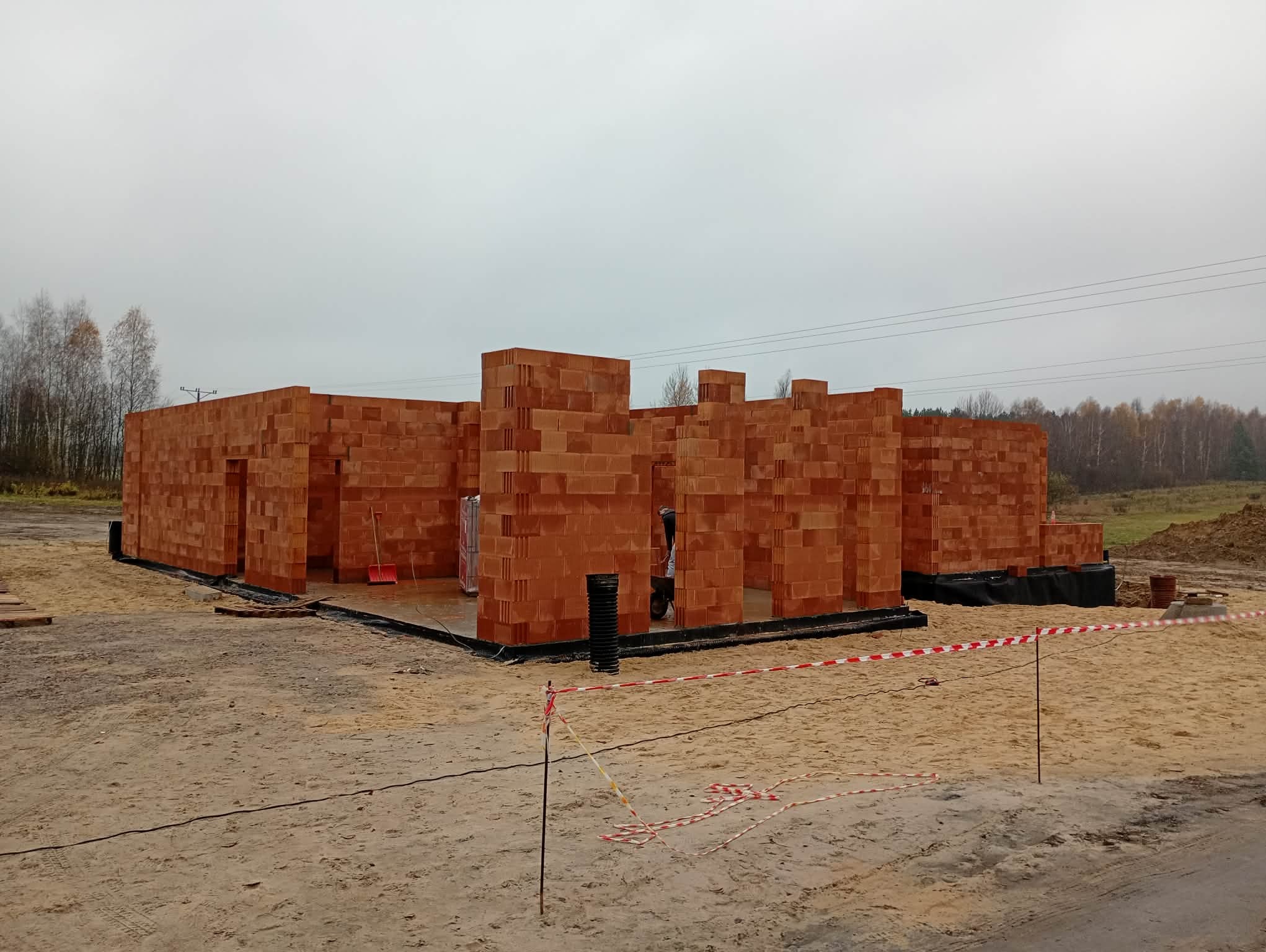 Wznoszenie ścian domu z czerwonej cegły ceramicznej. Widoczne fundamenty i izolacja. Na pierwszym planie piasek i taśma ostrzegawcza. Pochmurne niebo w tle.