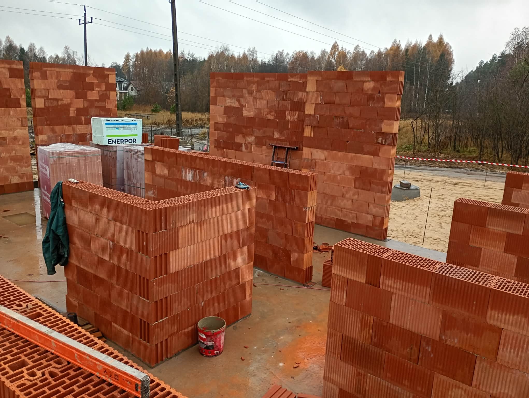 Wznoszenie ścian z czerwonej cegły ceramicznej na budowie domu jednorodzinnego. Widoczne bloczki Enerpor, poziomica i drabina. Jesienna sceneria w tle.