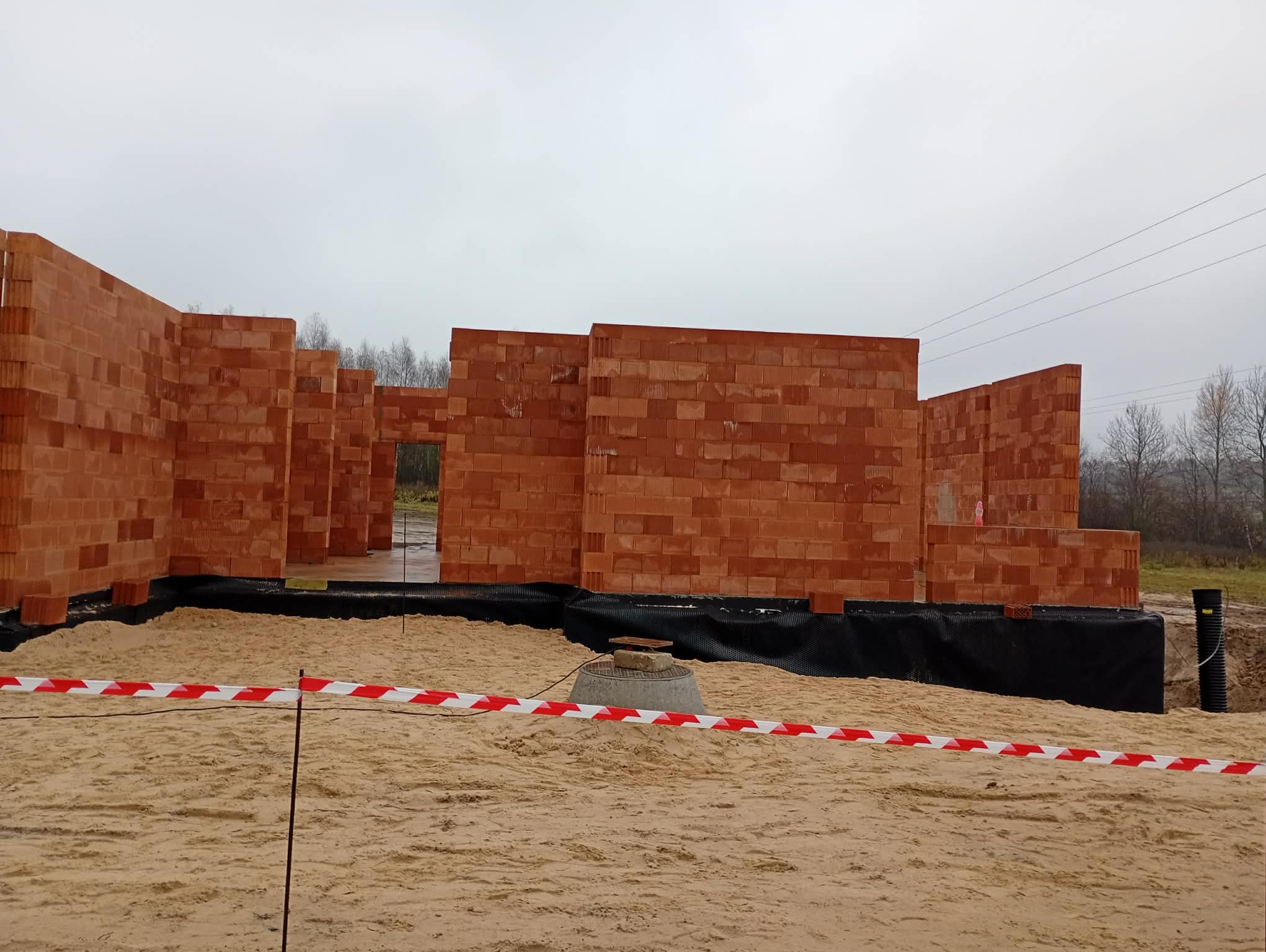 Wznoszone ściany z czerwonej cegły na fundamencie, zabezpieczone taśmą ostrzegawczą. Widoczne elementy instalacji. Pochmurne niebo w tle.