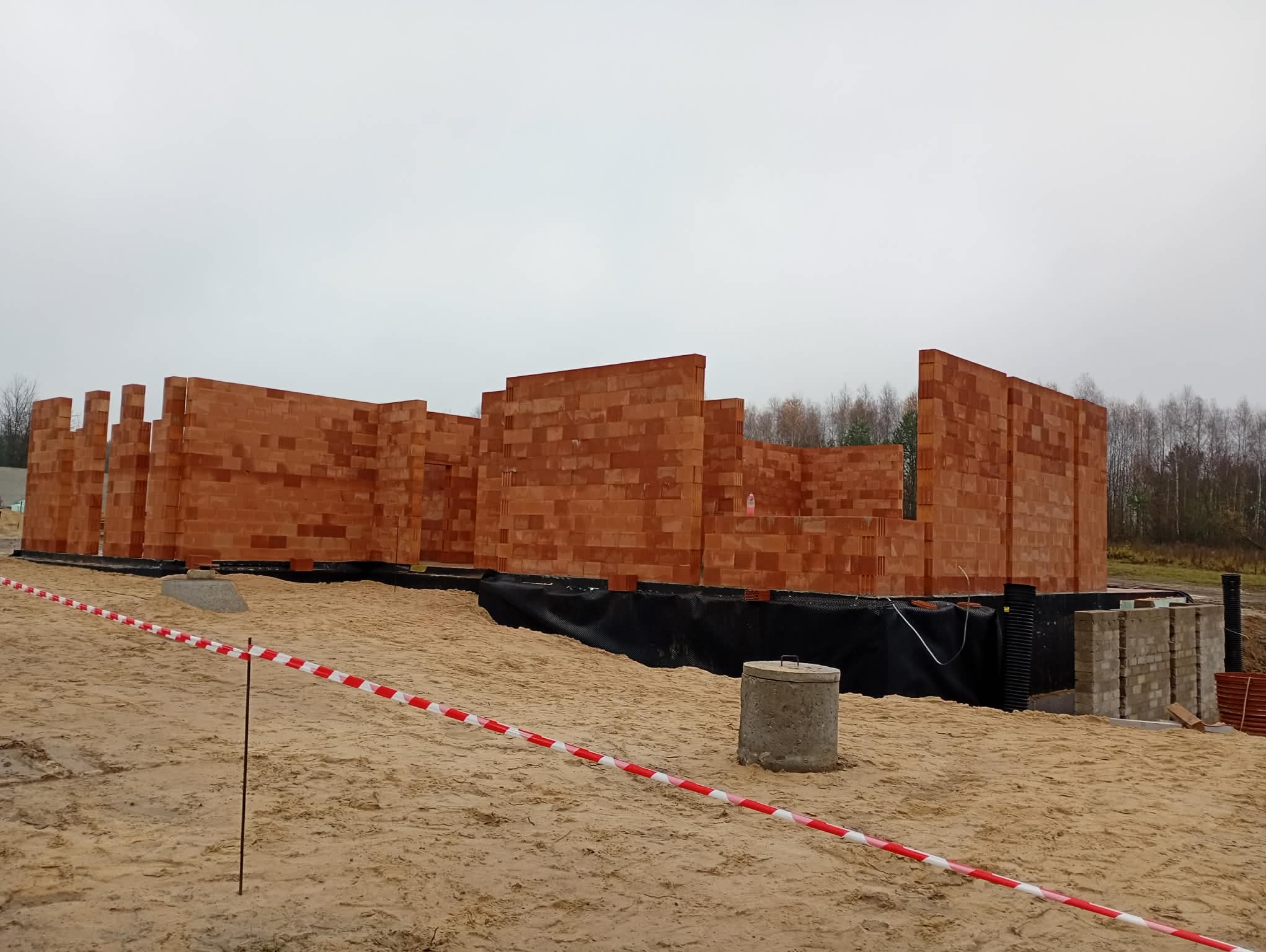 Wznoszenie ścian parteru z czerwonej cegły na fundamencie, zabezpieczonym czarną folią. Widoczne ogrodzenie terenu budowy taśmą ostrzegawczą.