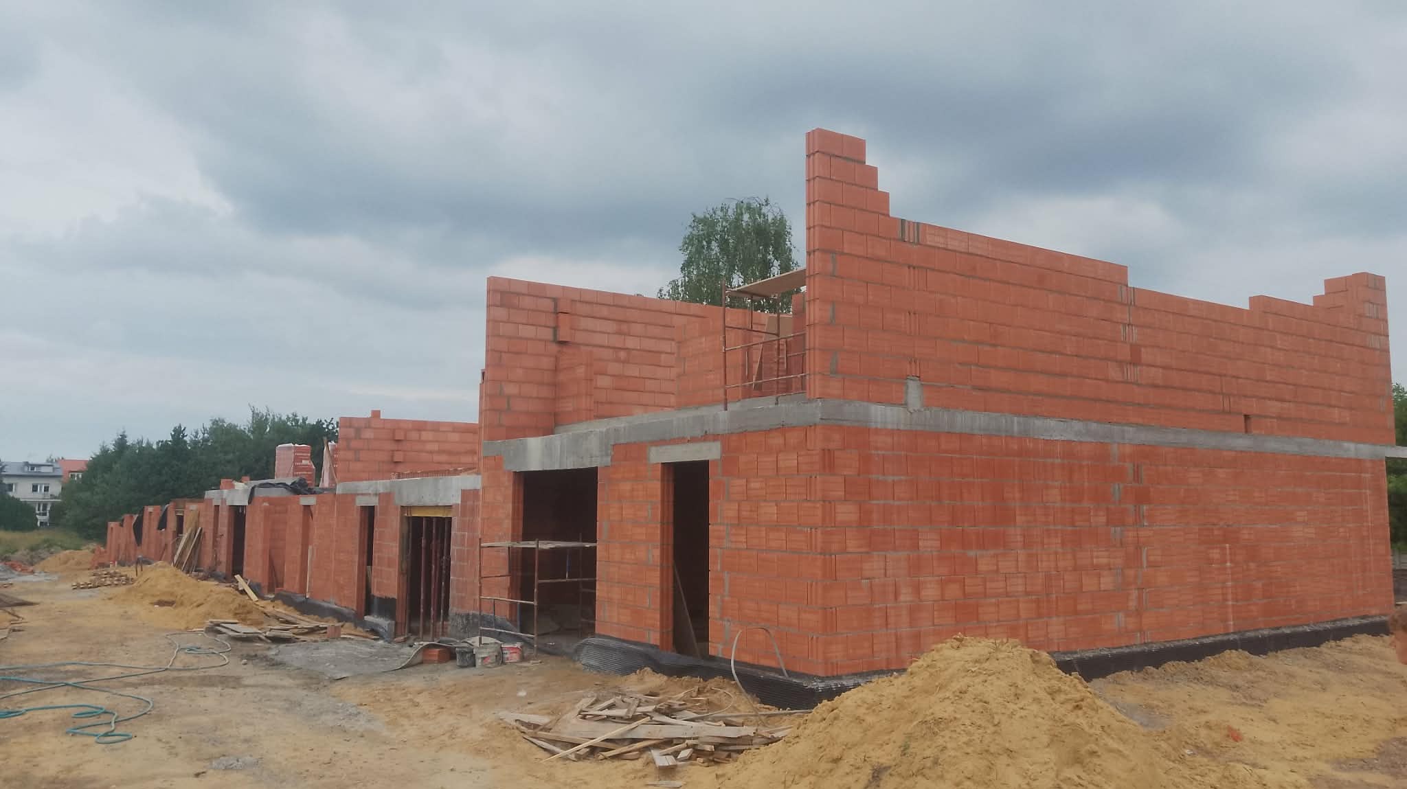 Wznoszenie ścian z czerwonej cegły na budowie szeregu domów jednorodzinnych. Widoczne rusztowania, piasek i materiały budowlane. Pochmurne niebo w tle.