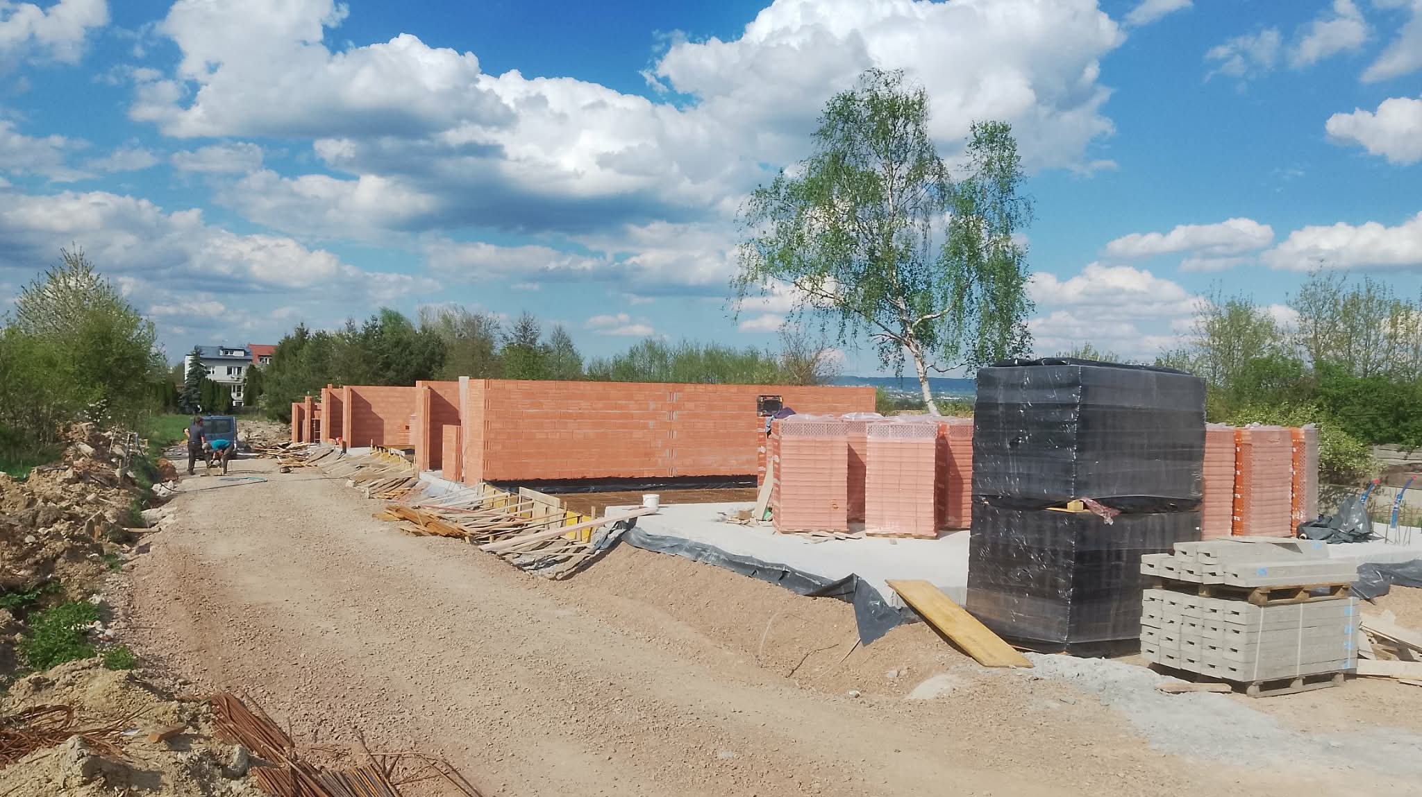 Budowa szeregówki z czerwonej cegły. Częściowo wzniesione ściany, palety z cegłami, teren budowy w słoneczny dzień. Widoczni pracownicy budowlani.