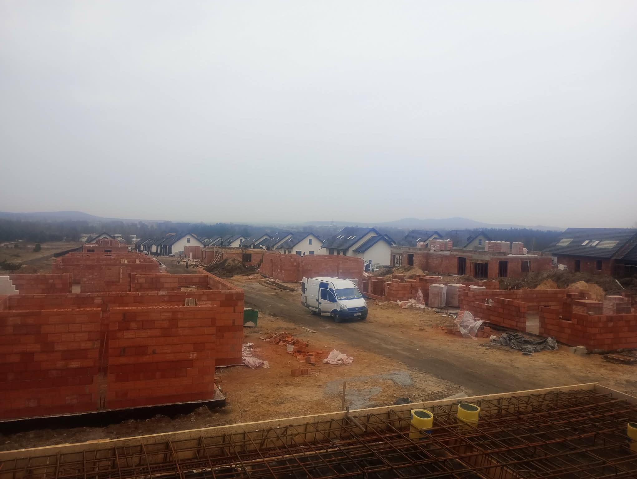 Budowa osiedla domów jednorodzinnych w trakcie wznoszenia ścian z czerwonej cegły. Na pierwszym planie zbrojenie, w tle biały bus na drodze dojazdowej.