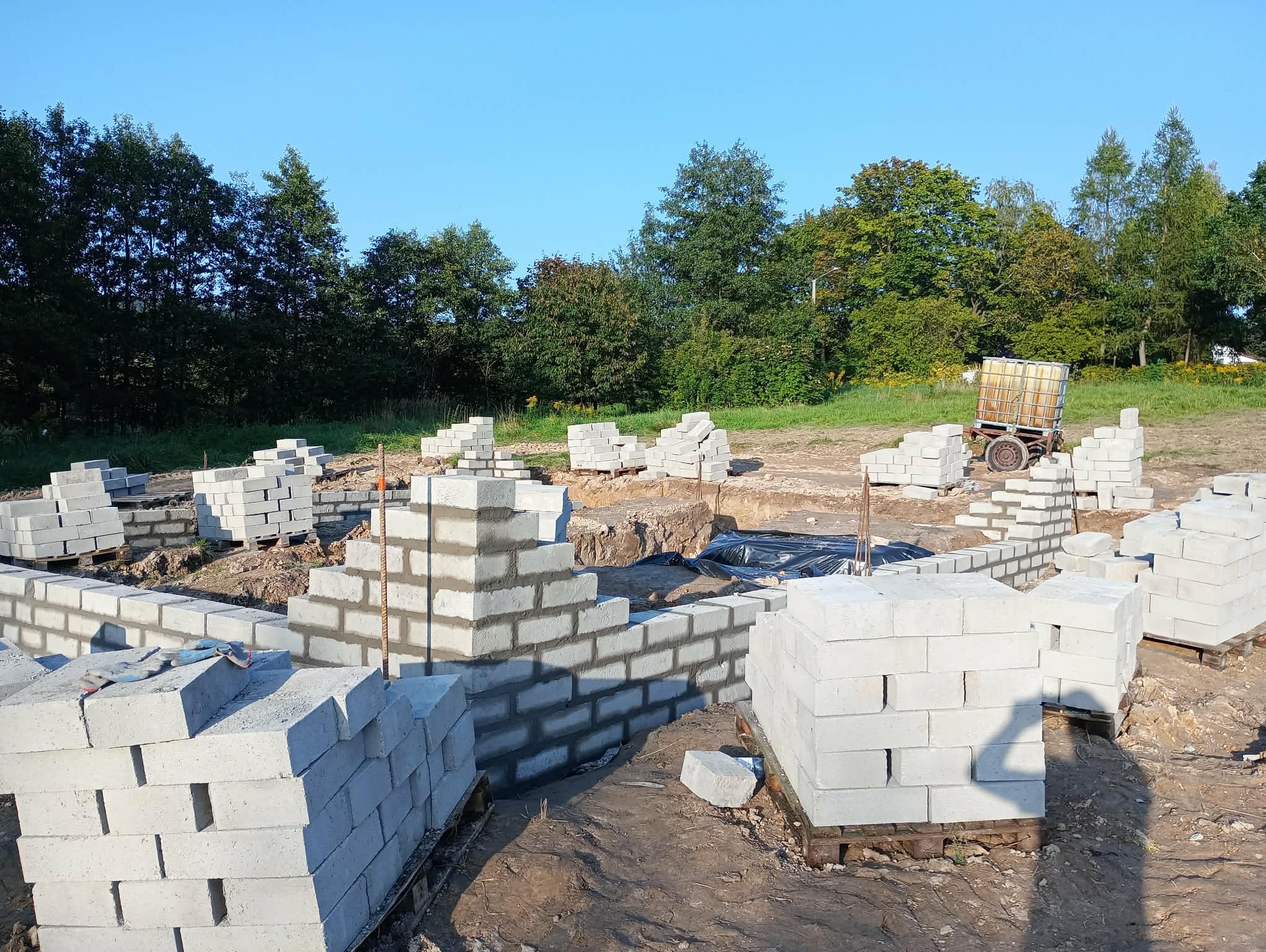 Wznoszenie ścian fundamentowych z bloczków betonowych na budowie domu jednorodzinnego. Widoczne zbrojenie i palety z bloczkami. W tle zieleń drzew i błękitne niebo.