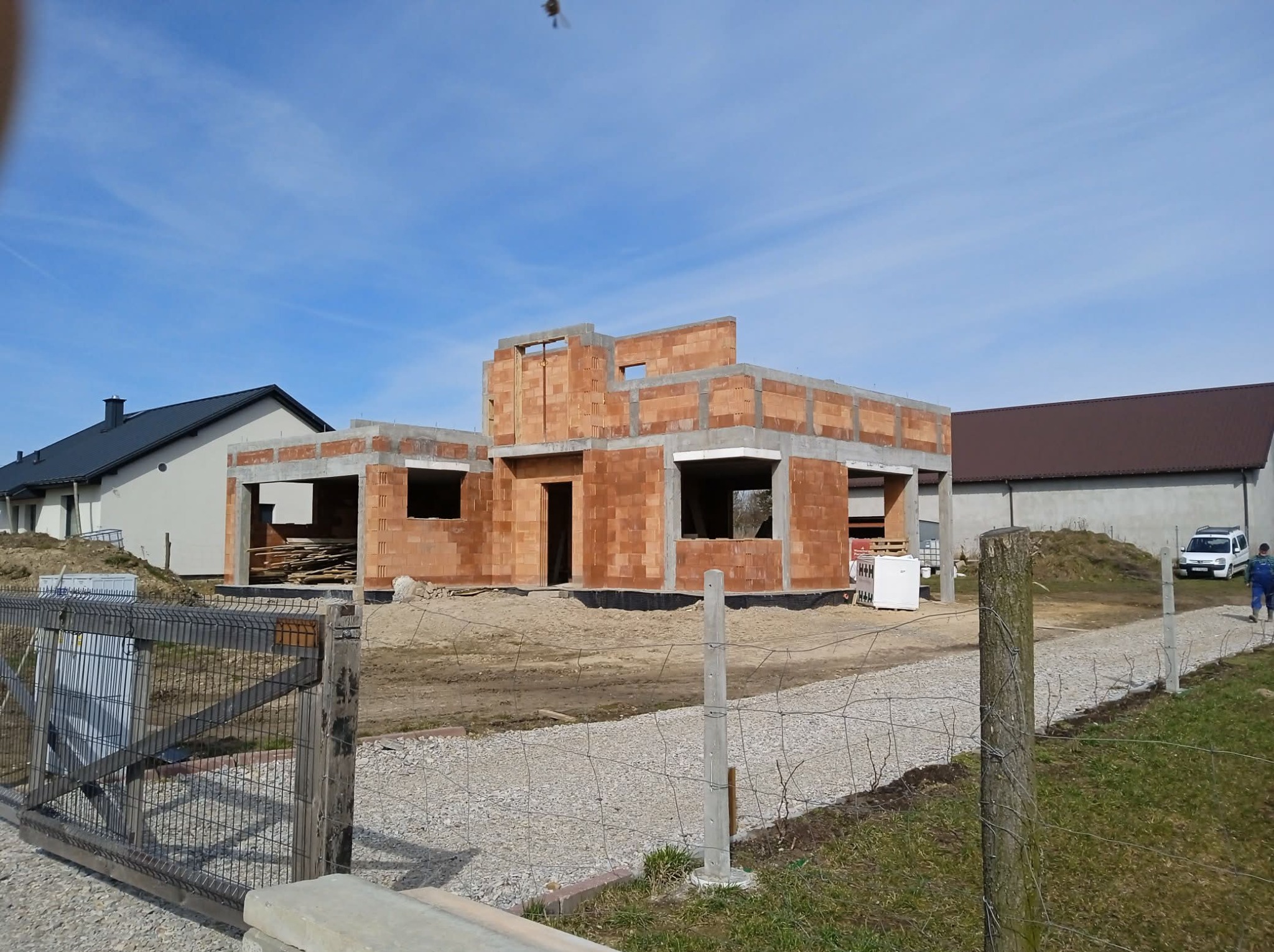 Budowa domu jednorodzinnego w stanie surowym otwartym, z widocznymi bloczkami ceramicznymi i betonowymi nadprożami, na tle błękitnego nieba i sąsiednich budynków.