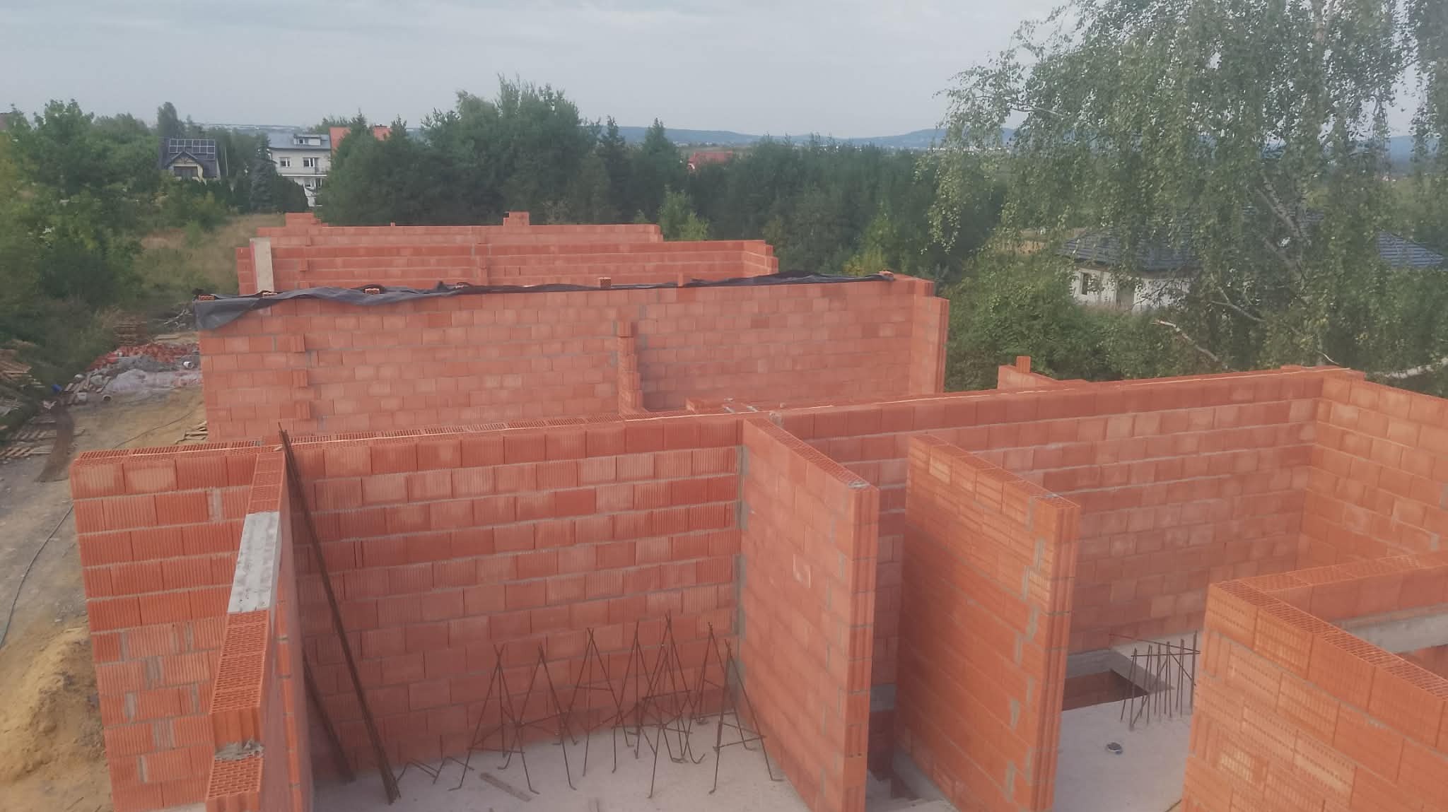 Wznoszenie ścian z czerwonej cegły dziurawki na budowie domu jednorodzinnego. Widok z góry na częściowo zrealizowane ściany działowe i obwodowe. W tle zieleń drzew i zabudowa.