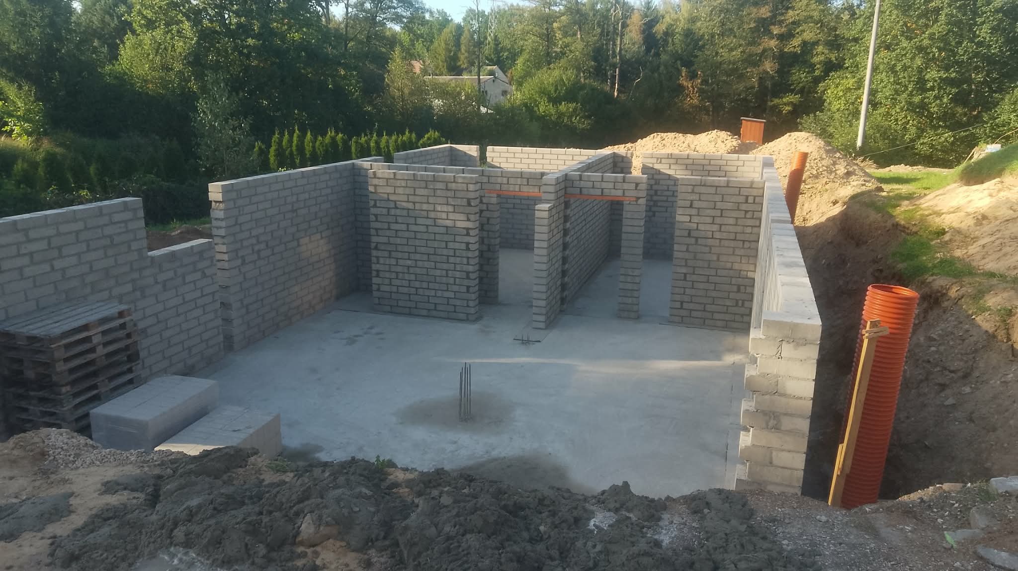 Wznoszenie ścian parteru z bloczków betonowych na fundamencie. Widoczne otwory drzwiowe i okienne, teren wokół przygotowany pod dalsze prace budowlane.
