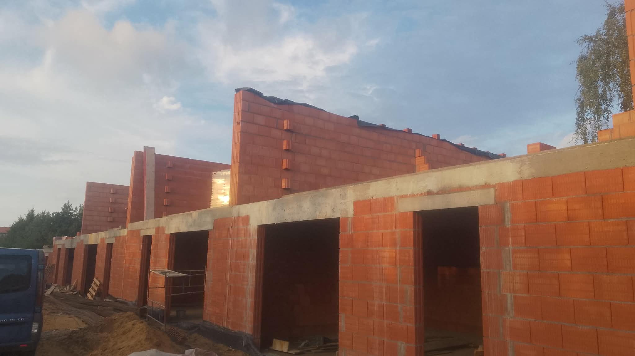 Wznoszenie ścian z czerwonej cegły na budowie szeregu budynków. Widoczne otwory okienne i drzwiowe, betonowe nadproża, oraz częściowo ułożona izolacja na górze muru.