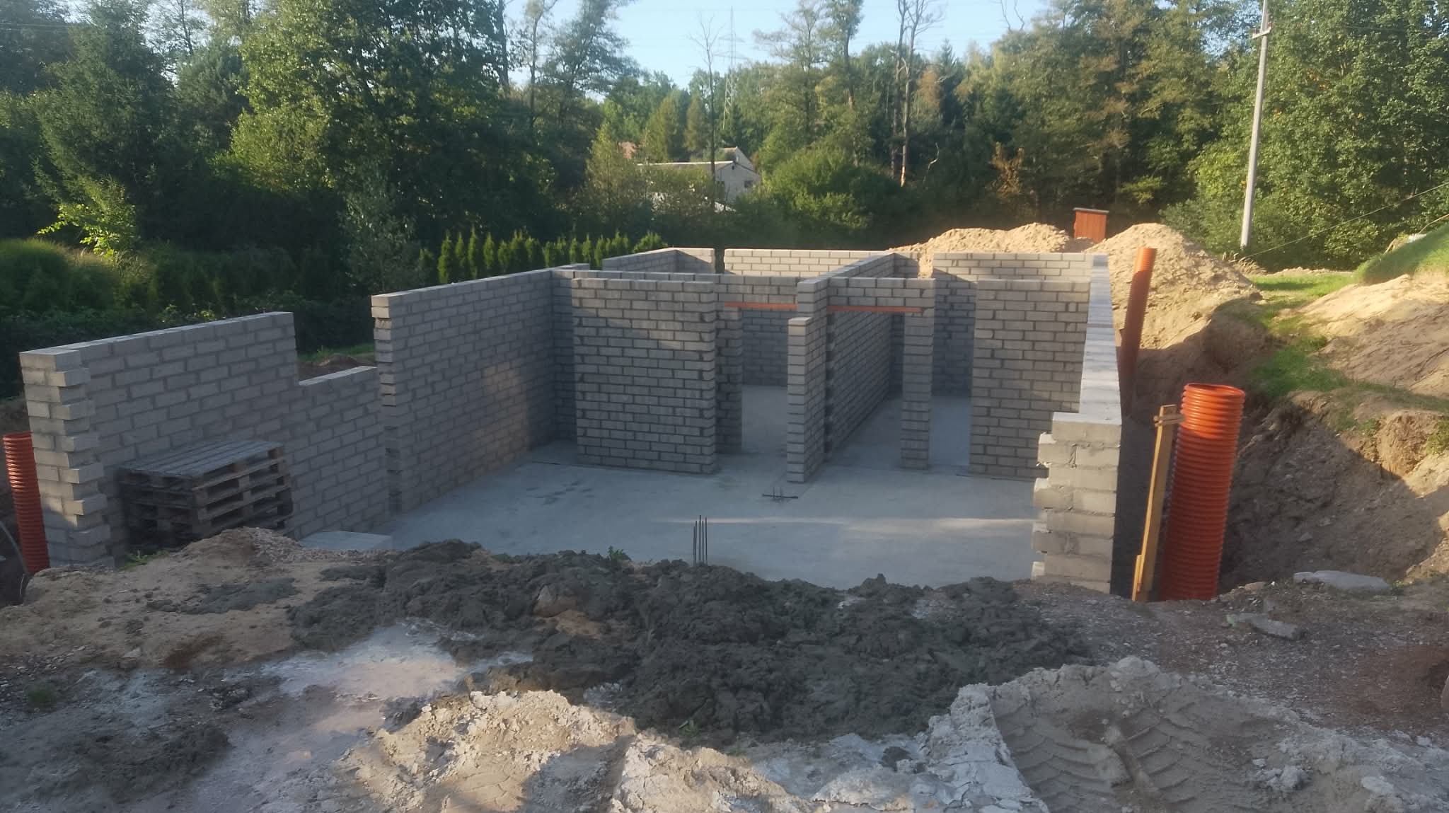 Wznoszenie ścian parteru z szarej cegły silikatowej na fundamencie. Widoczne pomarańczowe rury instalacyjne i paleta. Tło: drzewa i częściowo widoczny budynek.