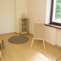 Jasne wnętrze sali zabaw z drewnianymi meblami, sztalugą z płótnem, dywanem i grami planszowymi na stoliku, widok na zieleń za oknem.