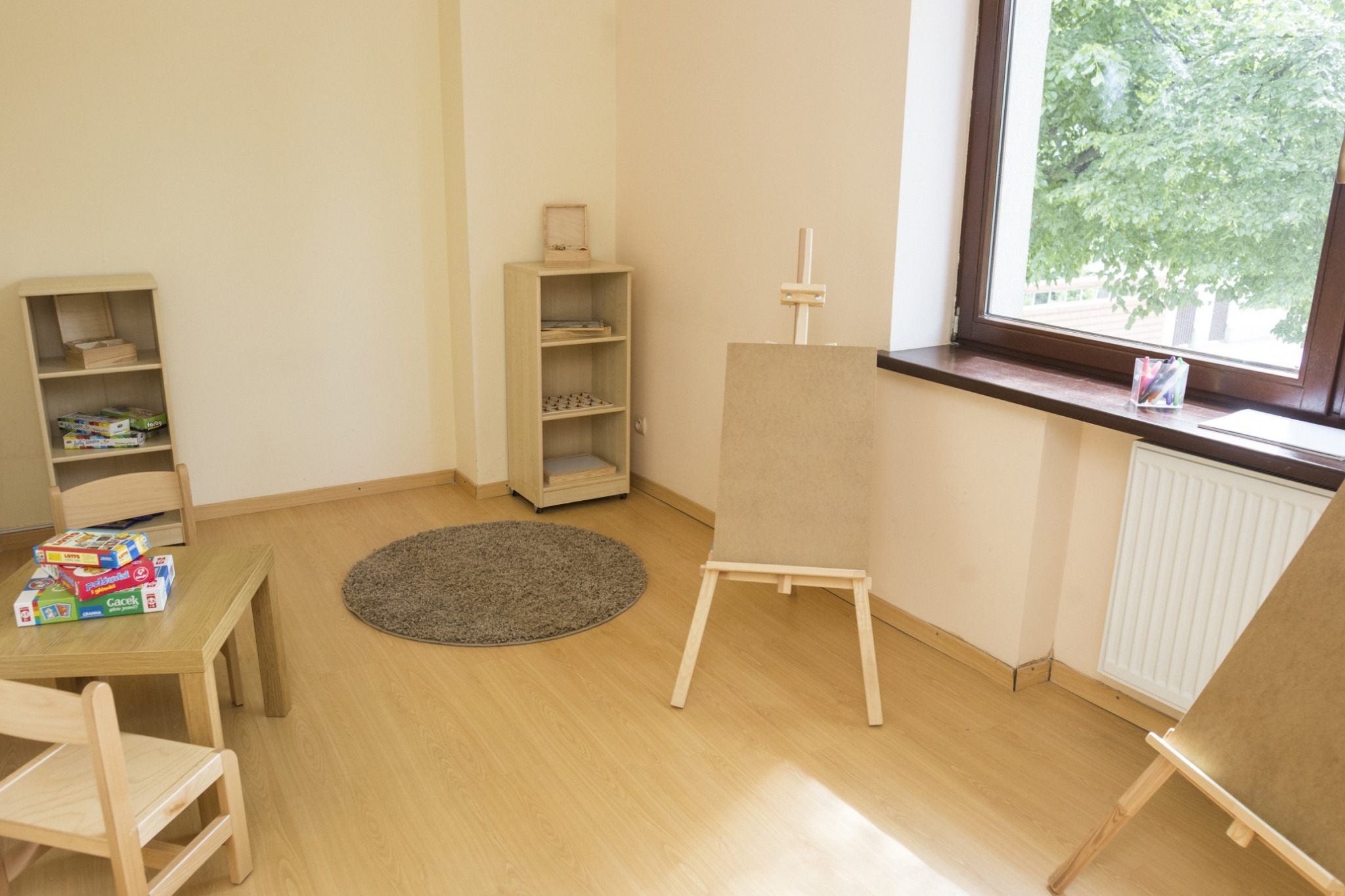 Jasne wnętrze sali zabaw z drewnianymi meblami, sztalugą z płótnem, dywanem i grami planszowymi na stoliku, widok na zieleń za oknem.
