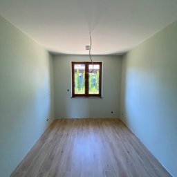 Prestige House Tomasz Caputa p - Pomalowane na jasno ściany i ułożona podłoga z paneli imitujących drewno w pustym pokoju z oknem. Widoczne kable od oświetlenia.