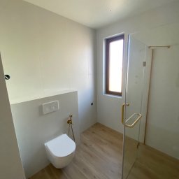 Prestige House Tomasz Caputa p - Elegancka łazienka z drewnopodobną podłogą, białymi płytkami ściennymi, podwieszaną toaletą i kabiną prysznicową ze złotymi detalami.