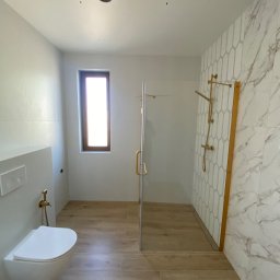 Prestige House Tomasz Caputa p - Nowoczesna łazienka z drewnopodobną podłogą, marmurowymi płytkami i kabiną prysznicową ze złotymi akcentami. Wisząca toaleta z bidetką. Okno z drewnianą ramą.