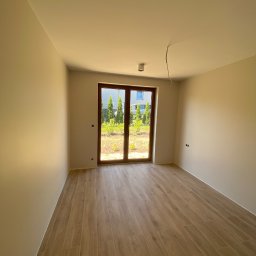 Prestige House Tomasz Caputa p - Jasne, puste pomieszczenie z drewnopodobną podłogą i widokiem na zieleń przez okno. Świeżo wykończone ściany i minimalistyczne oświetlenie sufitowe.