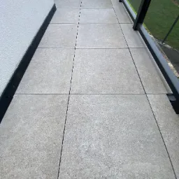 Wyłożony szarymi płytkami balkon z czarną balustradą i fragmentem ściany. Widok z góry, ukazujący regularny wzór płytek i minimalistyczny design balkonu.