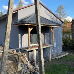 Nowo otynkowany dom jednorodzinny z drewnianą konstrukcją zadaszenia wejścia, widoczne betonowe słupy energetyczne i gruz budowlany.