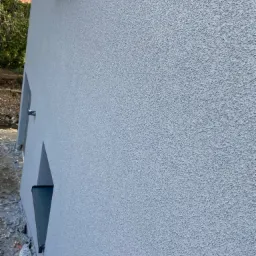 Ściana budynku z nową, jasnoszarą, ziarnistą strukturą, widoczne obramowania okien i fragment dachu w tle.