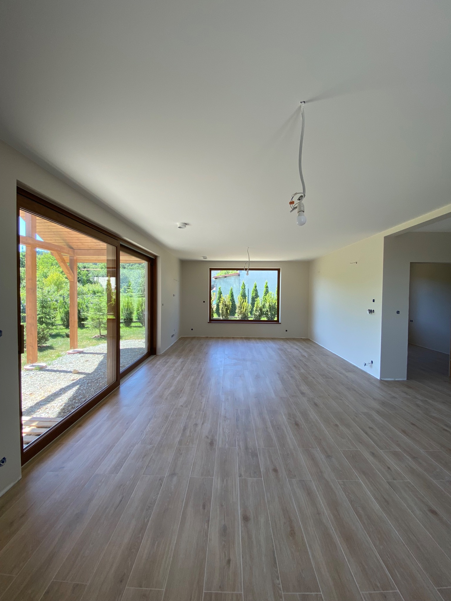 Jasne wnętrze z drewnopodobną podłogą i dużym oknem przesuwnym. Widoczne niedokończone gniazdka i wisząca żarówka. Za oknem zieleń i drewniana konstrukcja.