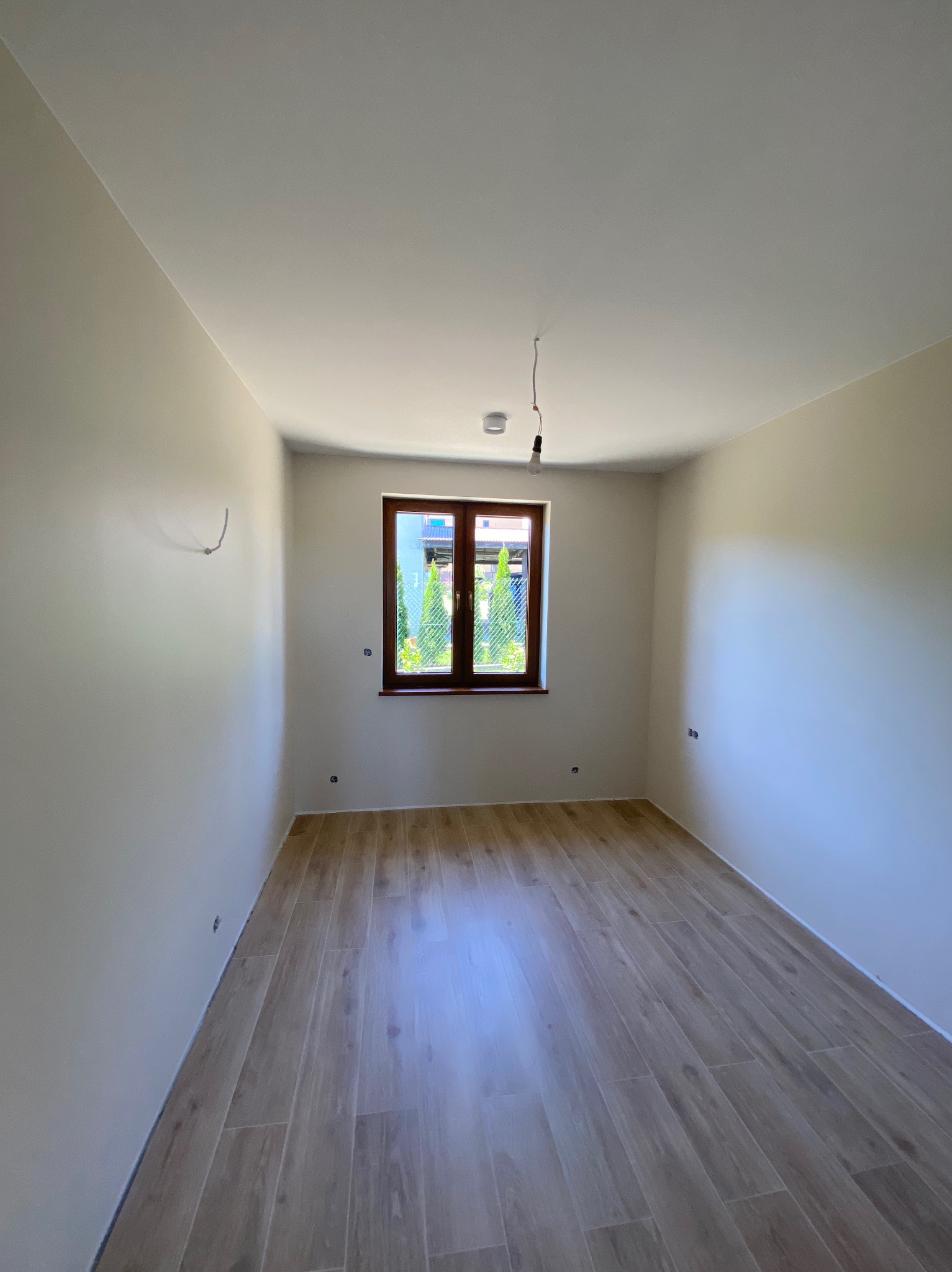 Jasne wnętrze po remoncie: pomalowane ściany w odcieniach beżu, drewniana podłoga, okno z brązową ramą. Widoczne wystające kable i żarówka.