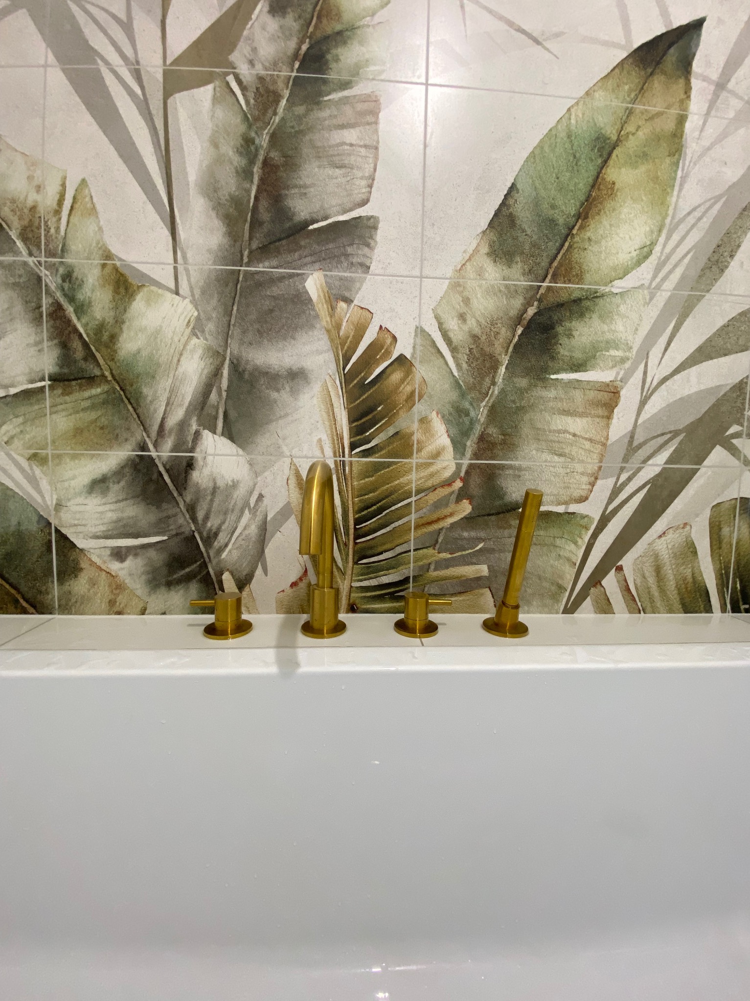 Biała wanna z złotą armaturą na tle ściany wyłożonej płytkami z motywem liści bananowca. Ujęcie centralne, prezentujące wykończenie łazienki.