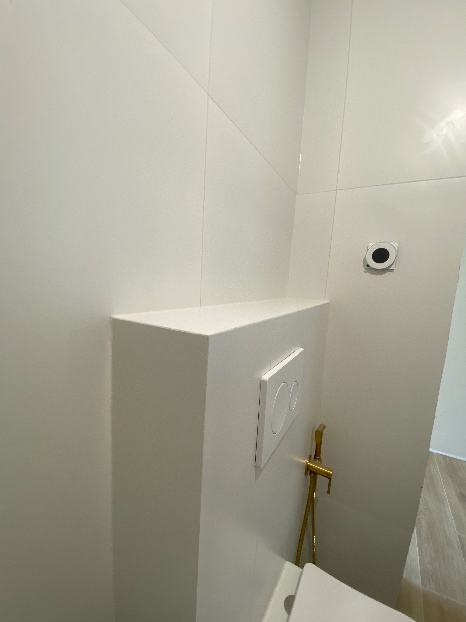 Biała, minimalistyczna łazienka z płytkami, widoczny fragment toalety, bidetta w kolorze złotym i przycisk spłuczki. Nowoczesne wykończenie.
