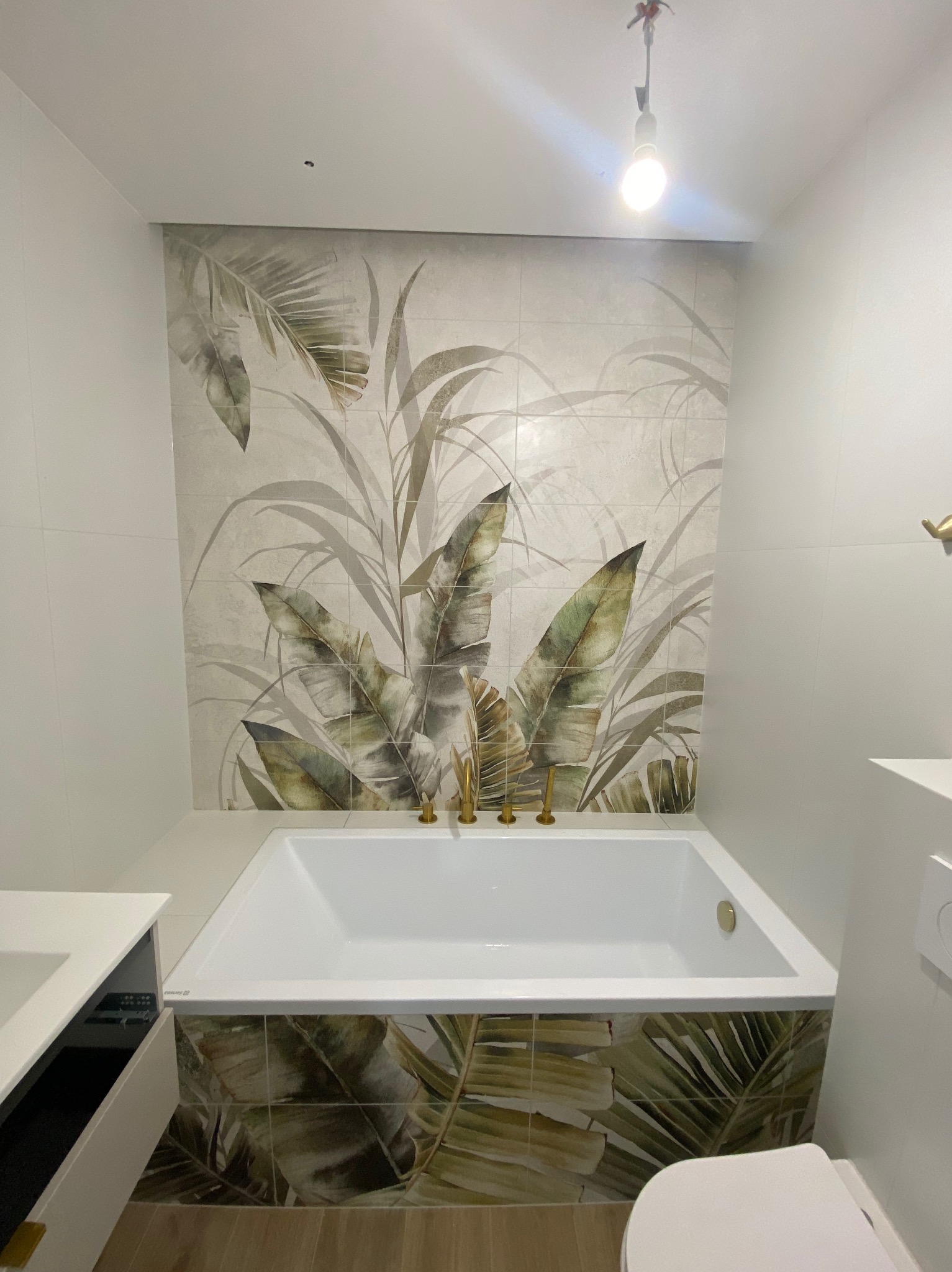 Wykończona łazienka z wanną obudowaną płytkami z motywem roślinnym, dopasowanymi do płytek na ścianie. Widoczne złote elementy armatury i biała ceramika.