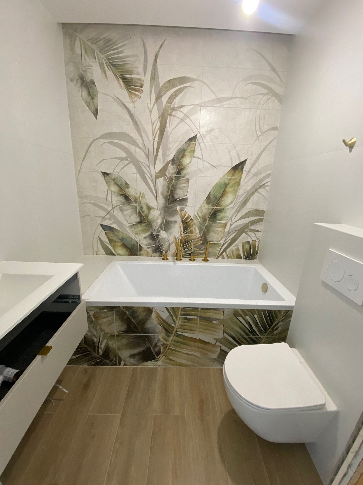 Nowoczesna łazienka z wanną obudowaną płytkami z motywem liści, drewnopodobna podłoga i biała armatura. Minimalistyczny design i naturalne akcenty.