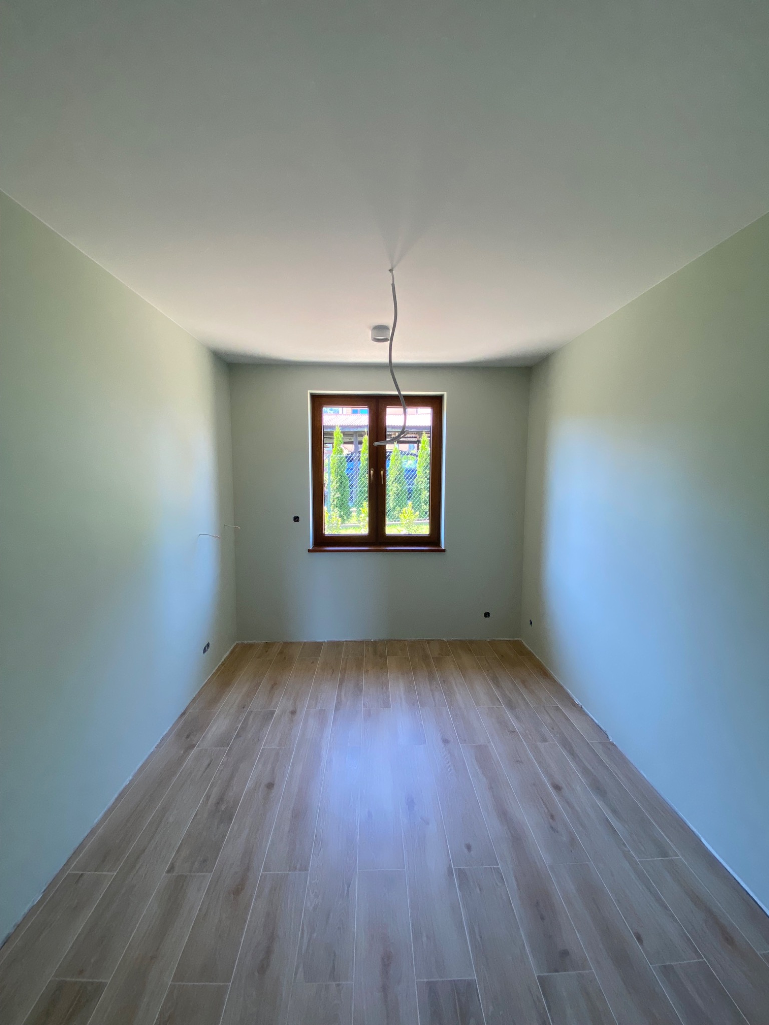 Pomalowane na jasno ściany i ułożona podłoga z paneli imitujących drewno w pustym pokoju z oknem. Widoczne kable od oświetlenia.