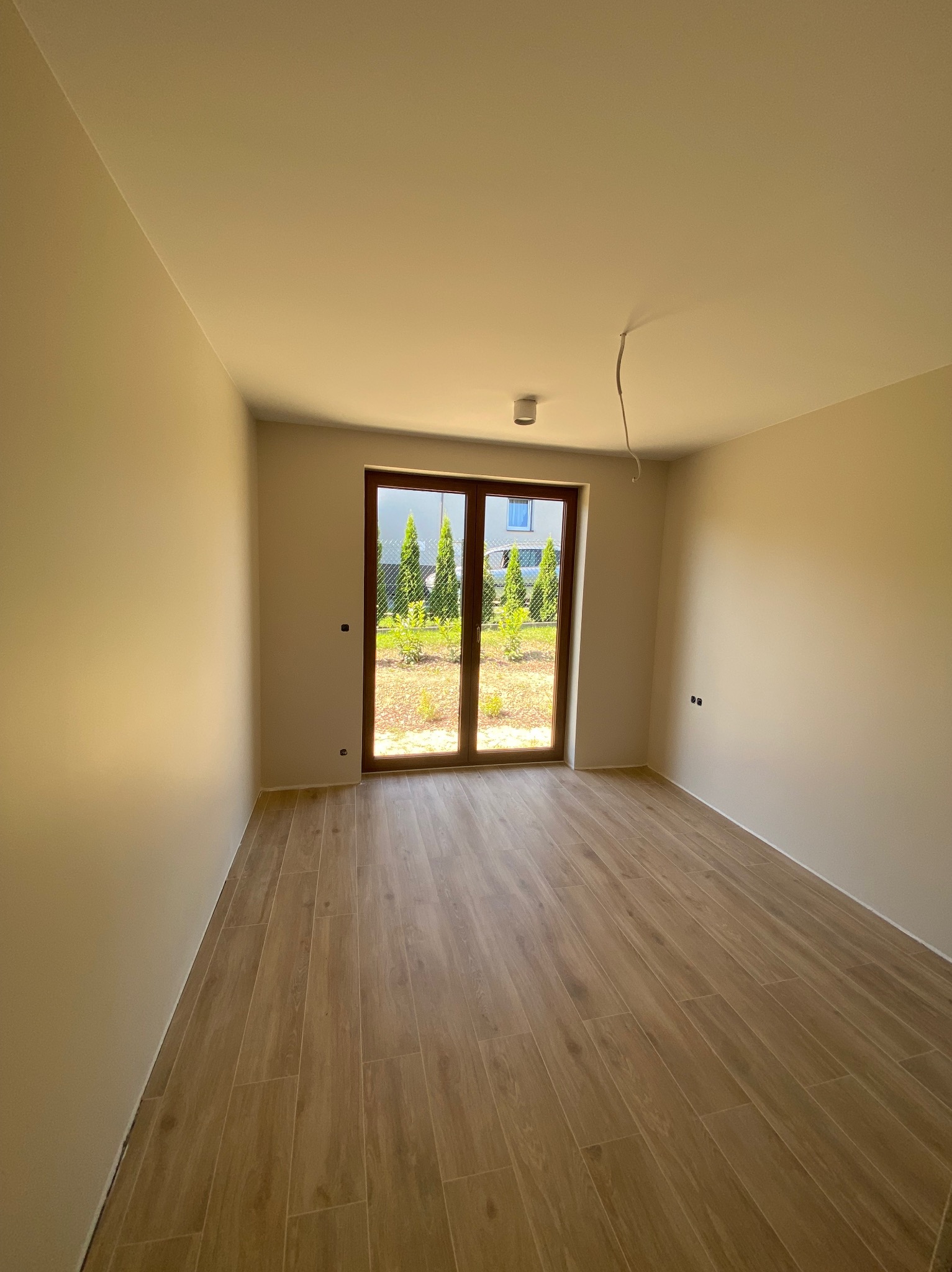 Jasne wnętrze pokoju z drewnopodobną podłogą i świeżo pomalowanymi ścianami. Widoczne okno balkonowe z widokiem na zieleń. Minimalistyczny design z surowym kablem.