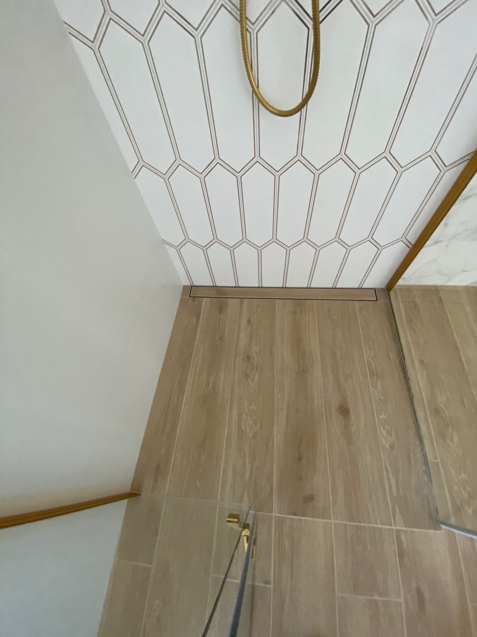 Wykończona łazienka z heksagonalnymi płytkami na suficie i drewnopodobną podłogą. Widoczny odpływ liniowy i złoty detal słuchawki prysznicowej.