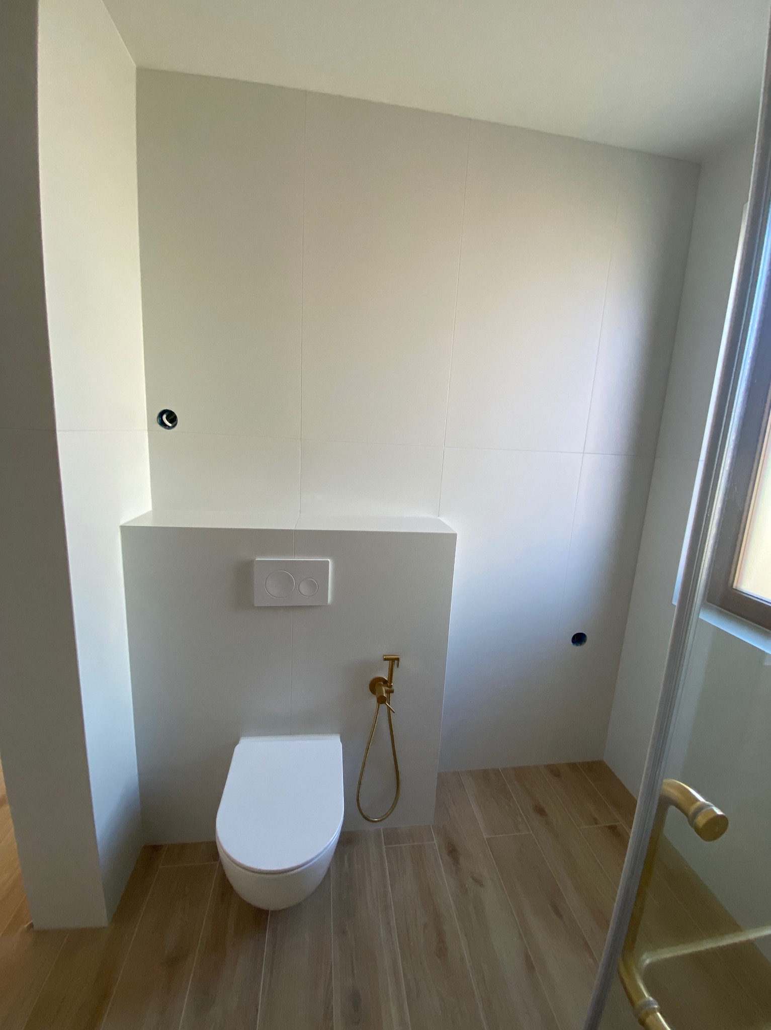 Minimalistyczna łazienka: białe płytki, podwieszana toaleta z złotą armaturą i drewnopodobna podłoga. Widoczne niedokończone otwory na armaturę.