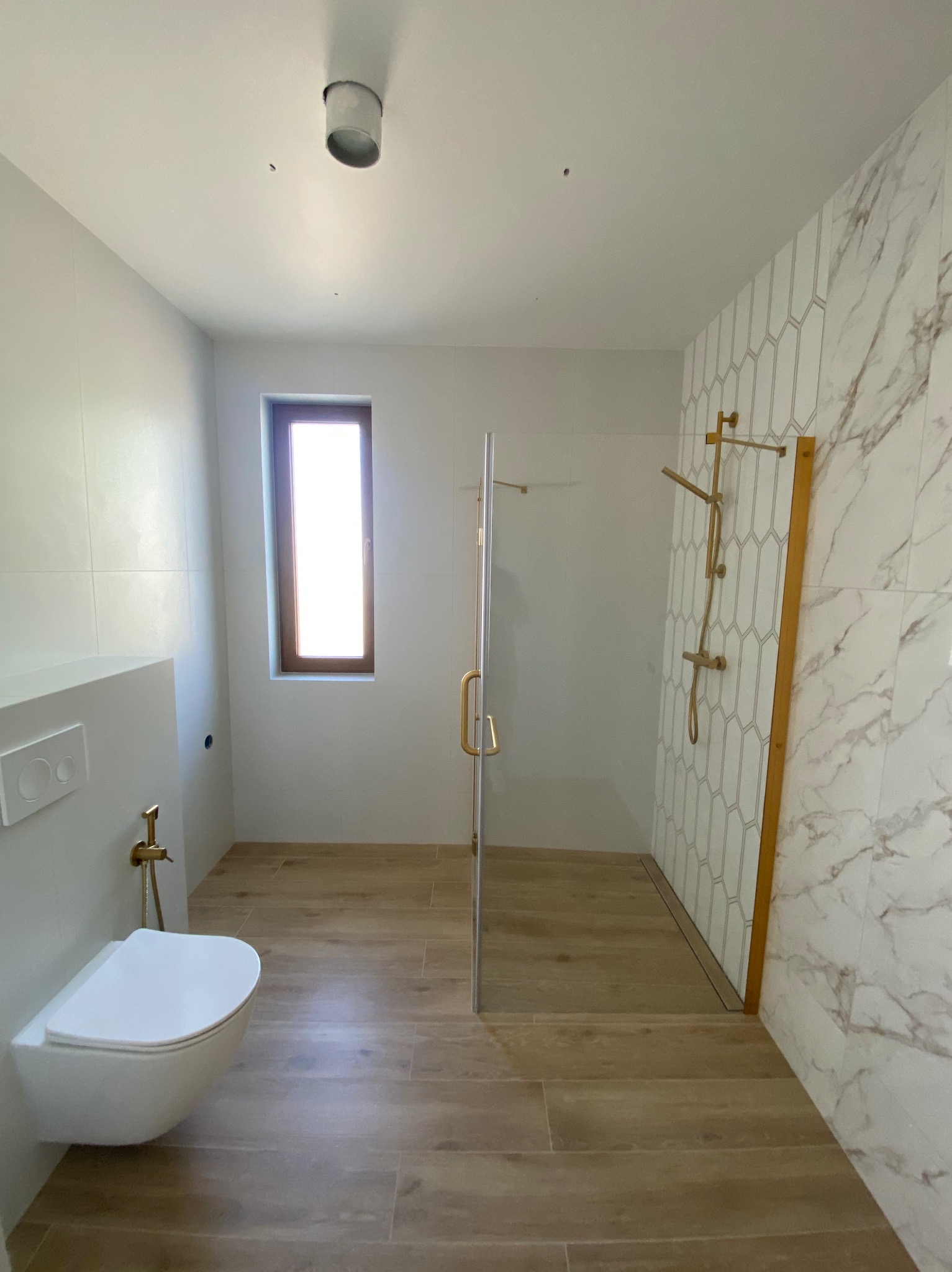 Nowoczesna łazienka z kabiną prysznicową typu walk-in, marmurowymi płytkami i drewnopodobną podłogą. Uwagę przyciąga złota armatura i minimalistyczna toaleta podwieszana.