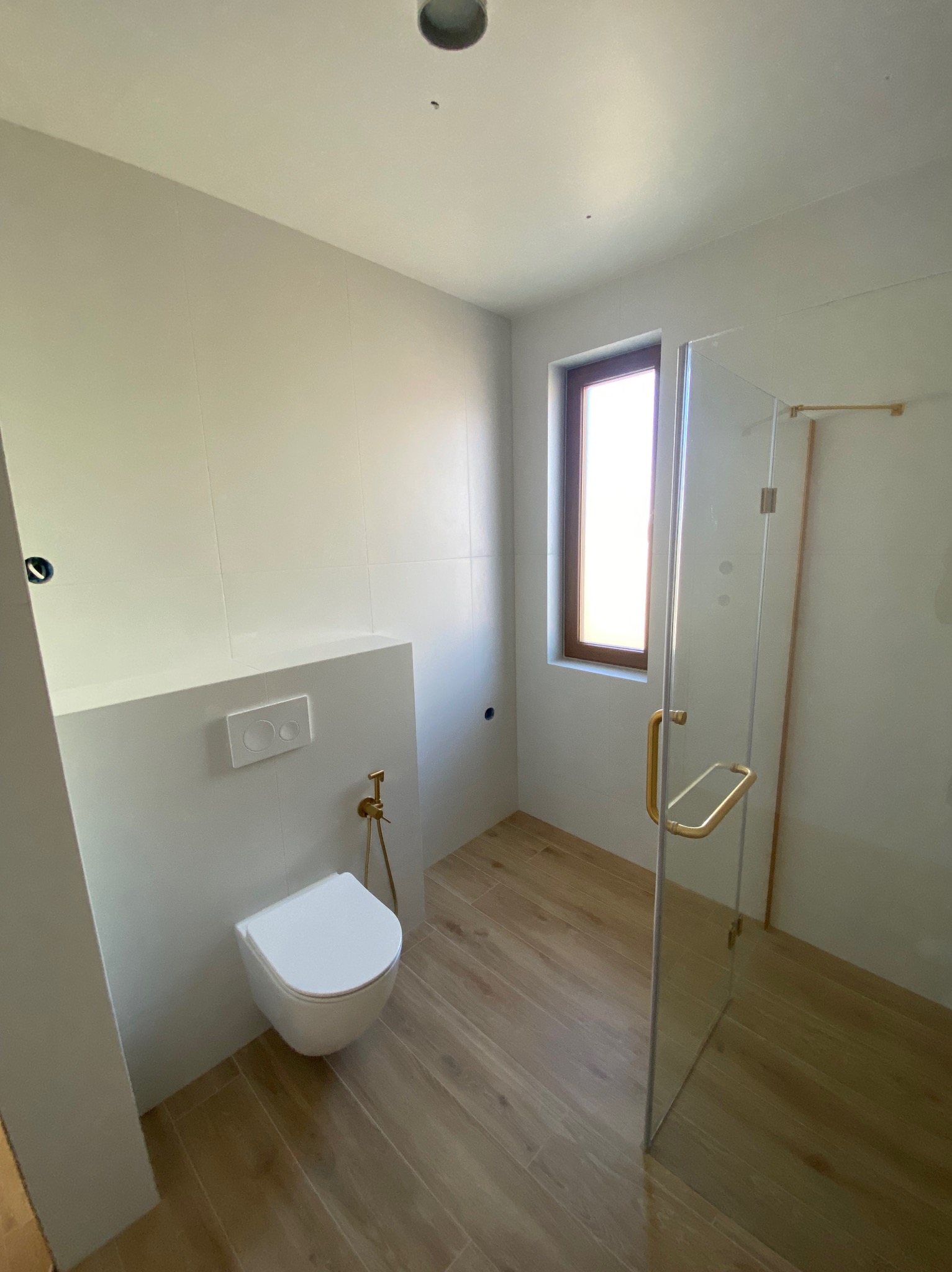 Elegancka łazienka z drewnopodobną podłogą, białymi płytkami ściennymi, podwieszaną toaletą i kabiną prysznicową ze złotymi detalami.