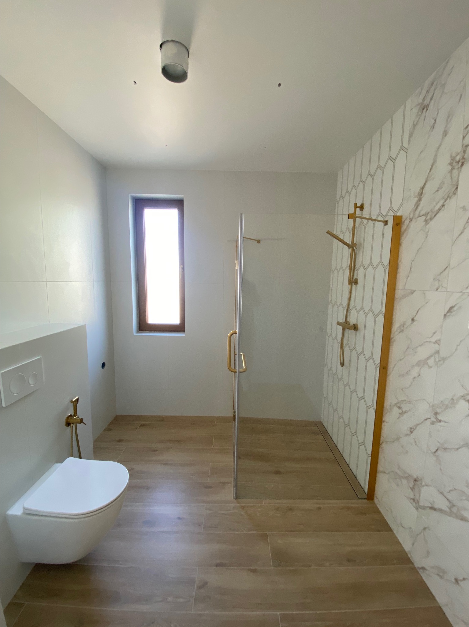 Nowoczesna łazienka z drewnopodobną podłogą, marmurowymi płytkami i kabiną prysznicową ze złotymi akcentami. Wisząca toaleta z bidetką. Okno z drewnianą ramą.