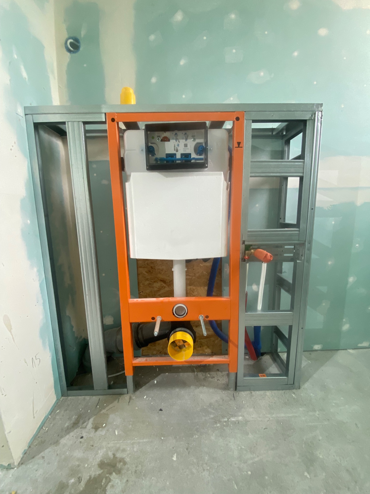 Stelaż podtynkowy WC z ramą w kolorze pomarańczowym, widoczny na tle ściany z płyt kartonowo-gipsowych. Ujęcie z bliska prezentuje detale instalacji wodno-kanalizacyjnej.