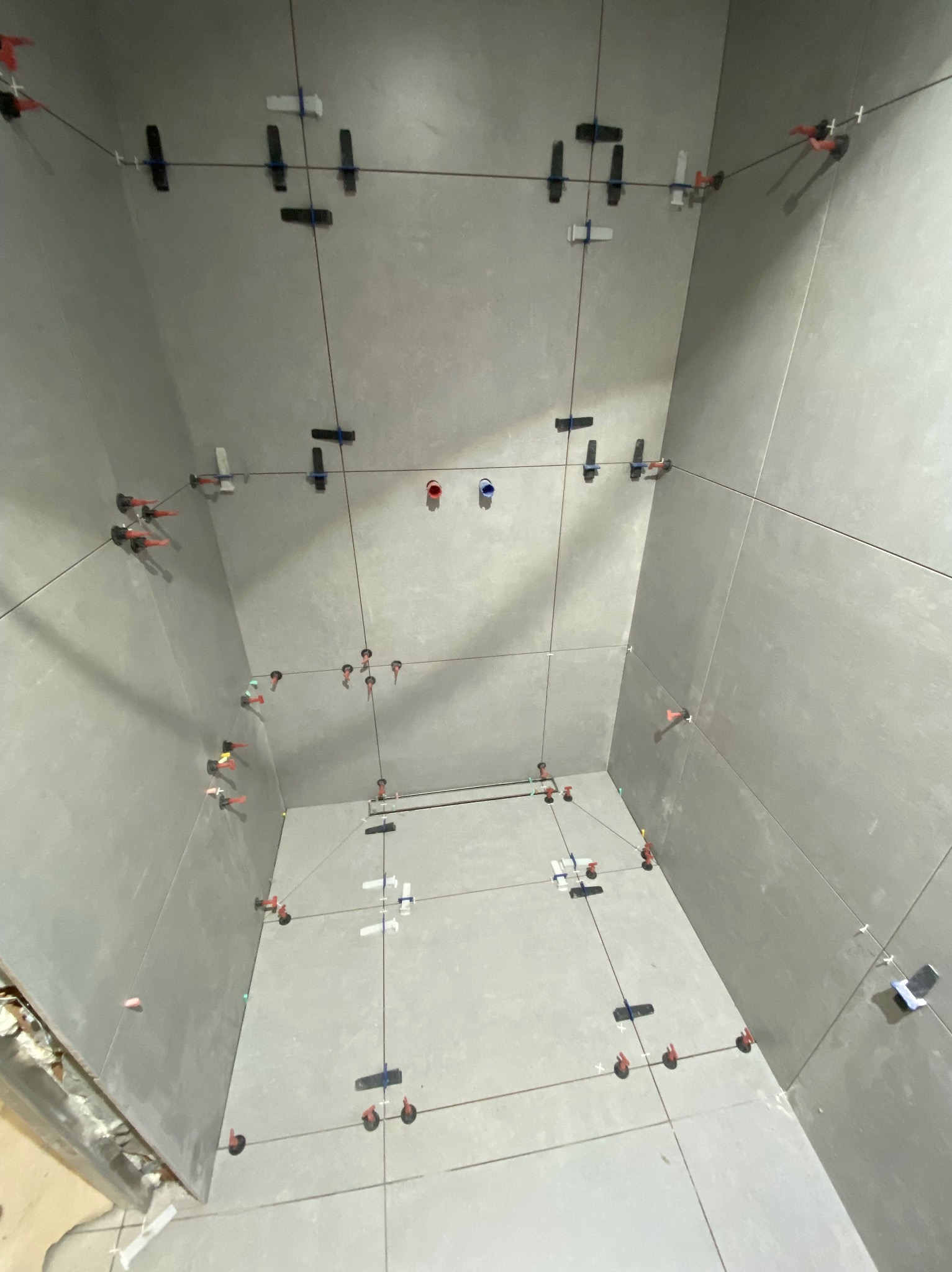 Szare płytki w trakcie montażu w narożniku, z użyciem systemu poziomowania płytek. Widoczne krzyżyki i kliny, oraz cienkie sznurki wyznaczające linie.