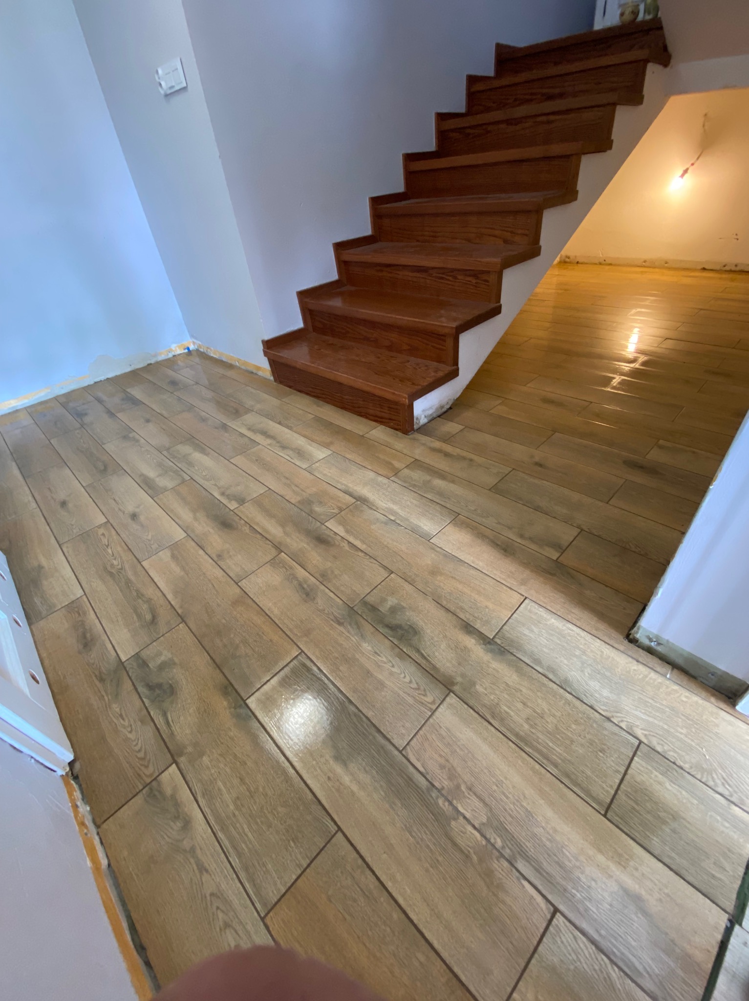 Ułożona jasnobrązowa podłoga imitująca drewno prowadząca do drewnianych schodów w jasnym pomieszczeniu. Widoczne niedokończone wykończenia przy ścianach.
