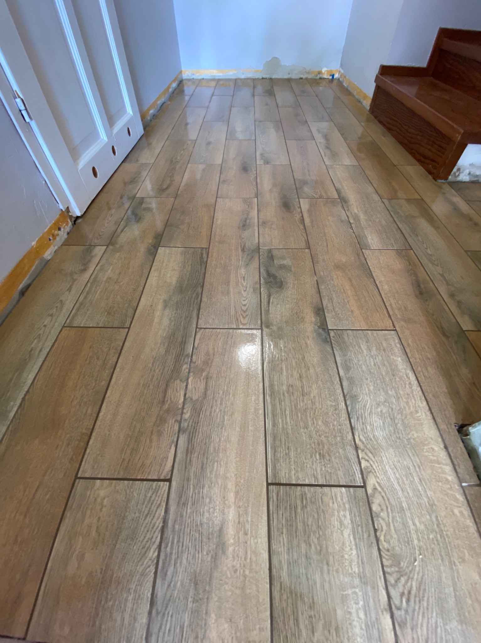 Ułożone płytki podłogowe imitujące drewno w korytarzu, widoczne schody i fragment drzwi. Wykończenie podłogi w odcieniach brązu i beżu.