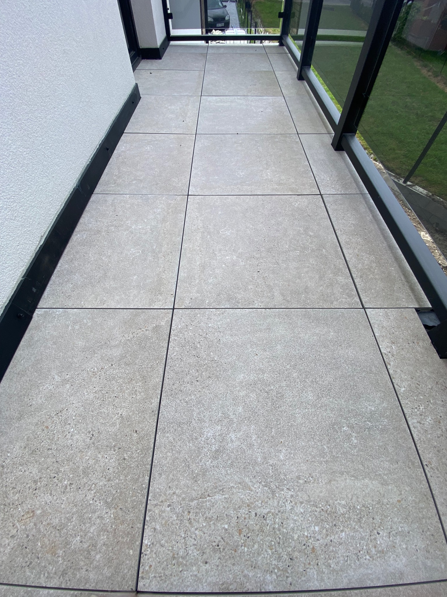 Wyłożony szarymi płytkami balkon z czarną balustradą i fragmentem ściany. Widok z góry, ukazujący regularny wzór płytek i minimalistyczny design balkonu.