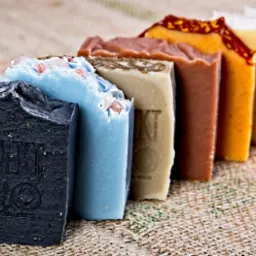 Ręcznie robione kostki mydła o różnych kolorach i fakturach, ułożone w rzędzie na jutowym tle, pierwsze mydło z napisem 'PROJK MOLO'.